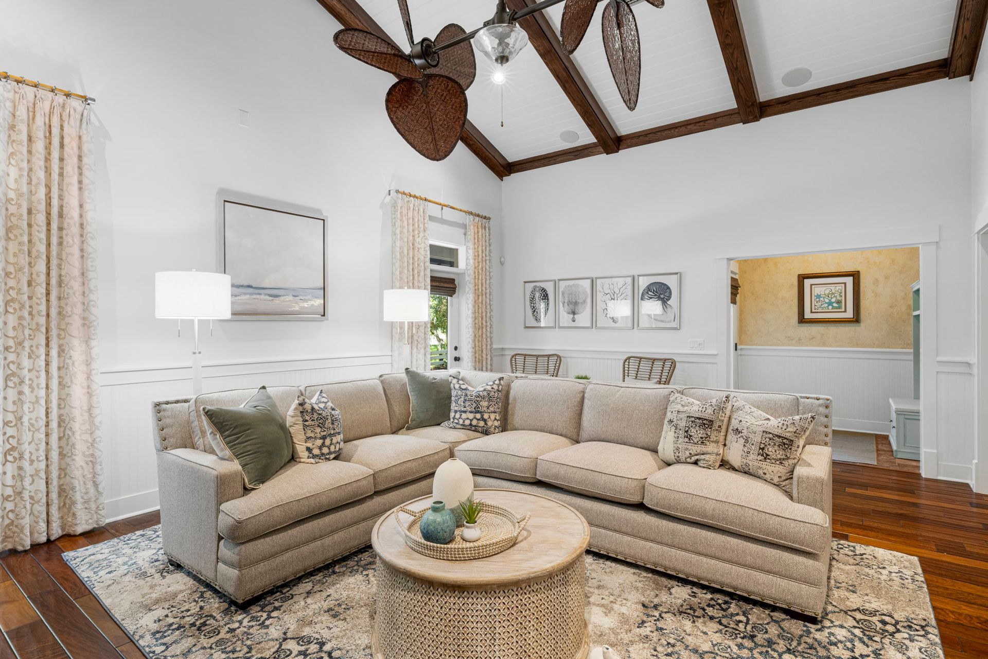 Cozy living room with beige sectional, patterned rug, floor lamps, and wood-beamed ceiling with ceiling fan.
