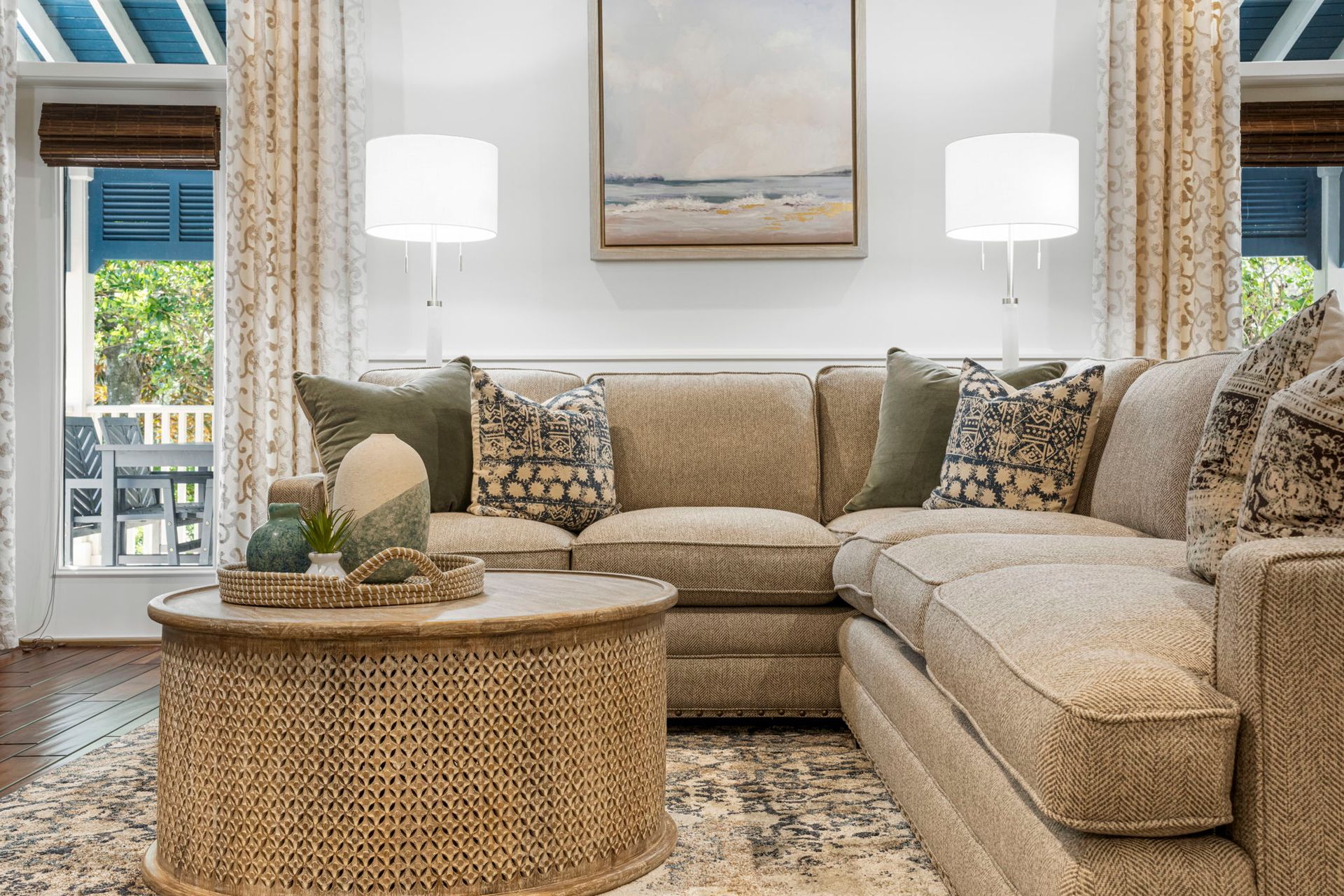 Bright living room with beige sectional sofa, woven coffee table, lamps, and large windows