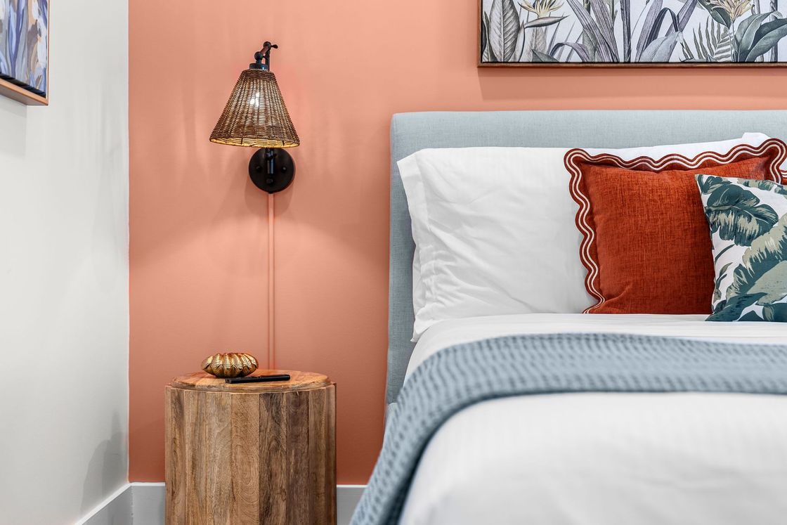 Peach bedroom wall with bedside lamp, tree-stump table, and bed with white bedding and orange pillow