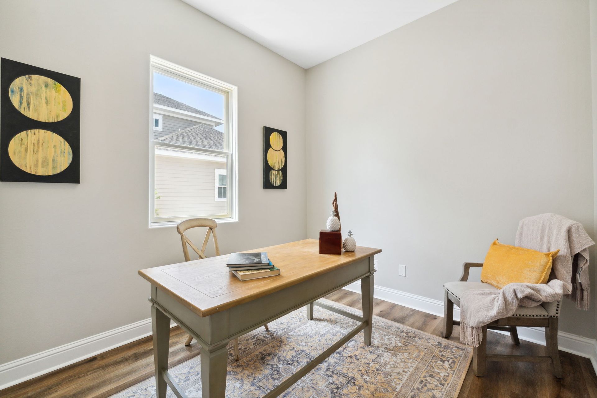 Bright home office with wooden desk, chair, rug, wall art, and a cushioned armchair by the window