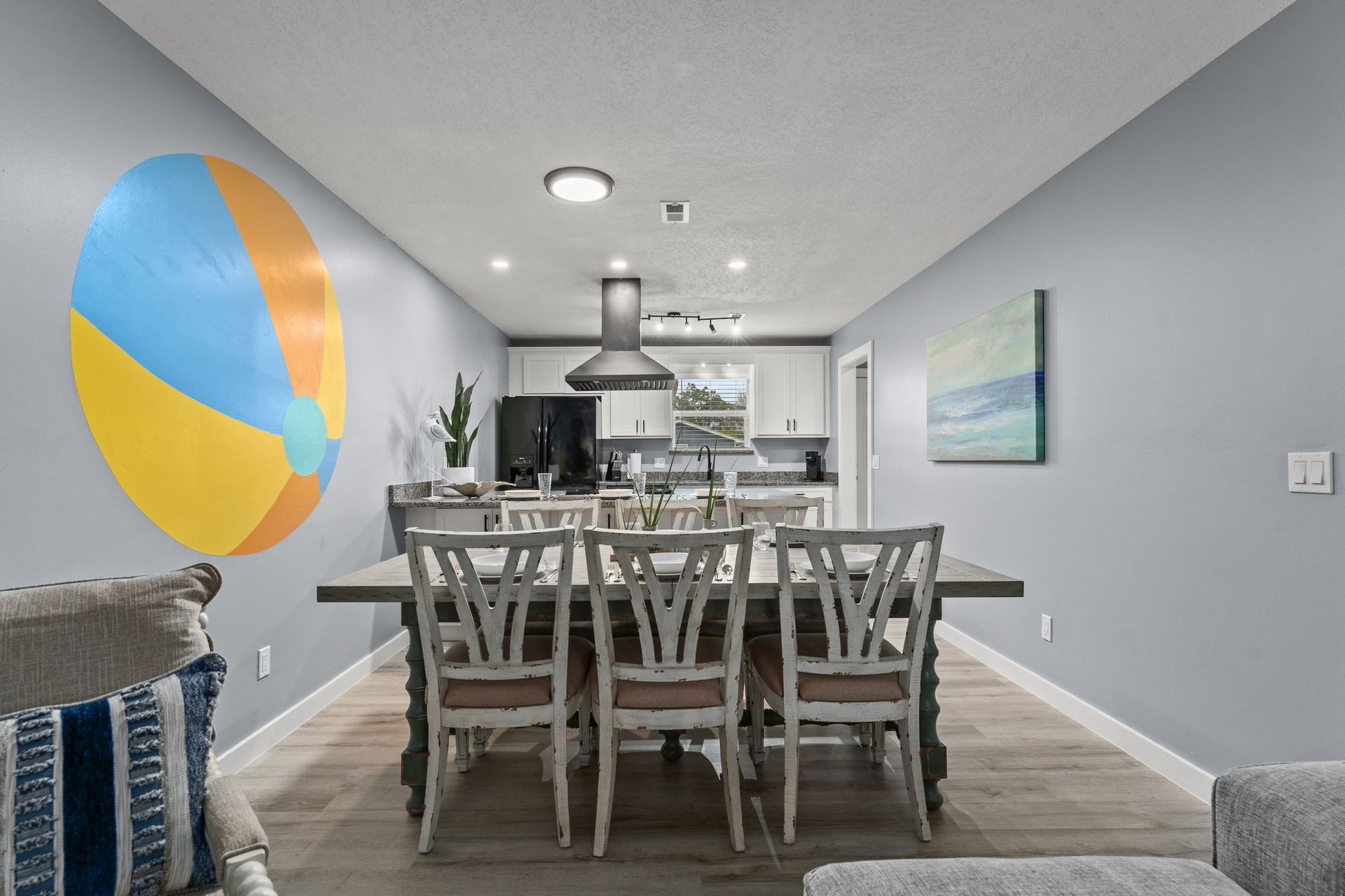 Modern dining room with gray table, six chairs, blue accent wall art, and open kitchen in background