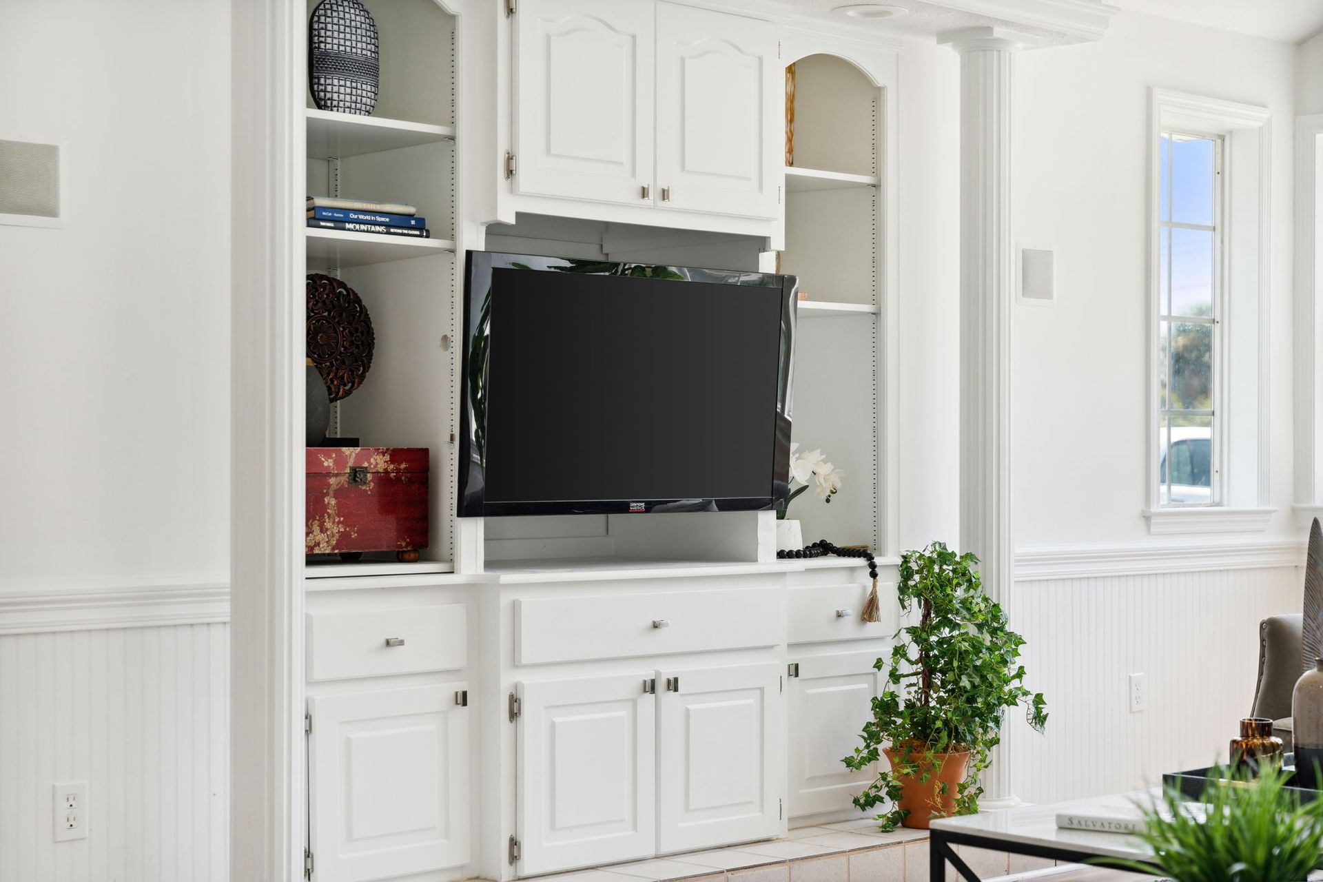White built-in living room cabinets with a TV, shelves, and a potted plant by a bright window