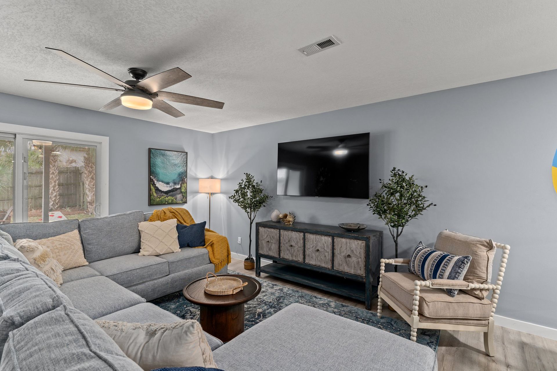 Bright living room with gray sectional, wall-mounted TV, ceiling fan, and woven chair by the window.
