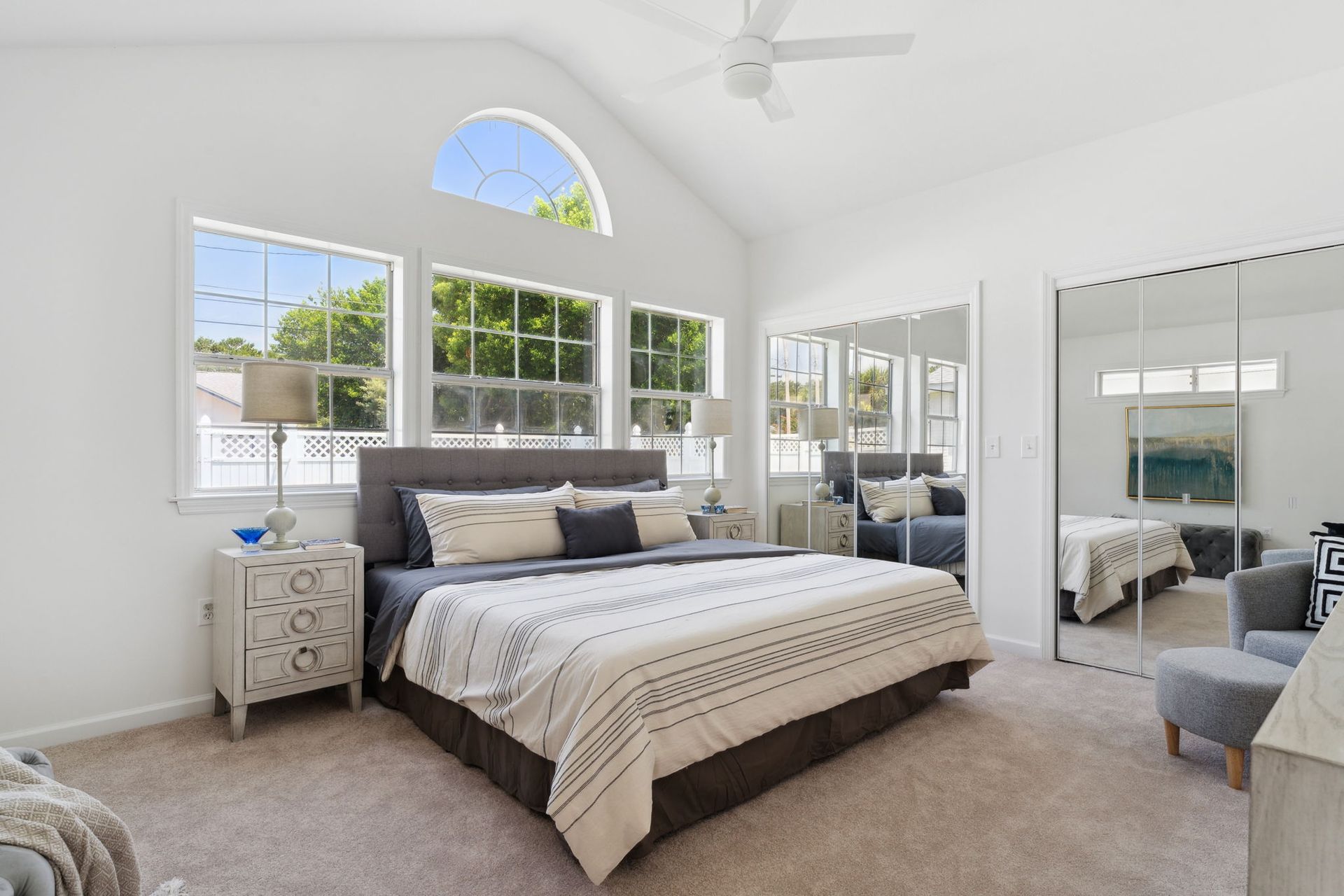 Bright bedroom with a large bed, gray accents, and lots of natural light from windows and a mirrored closet.