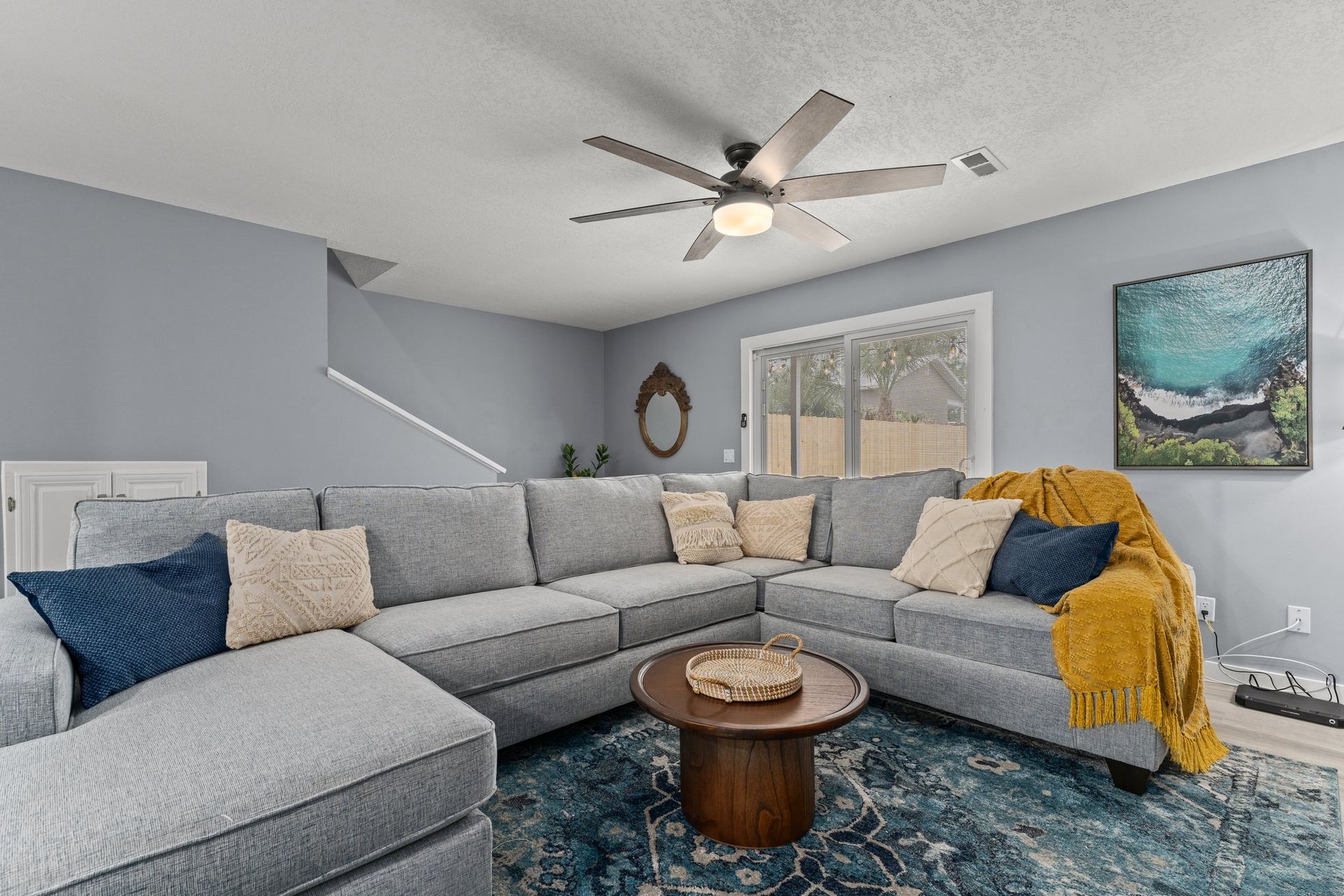 Gray sectional sofa in a bright living room with blue rug, ceiling fan, wall art, and sliding glass doors