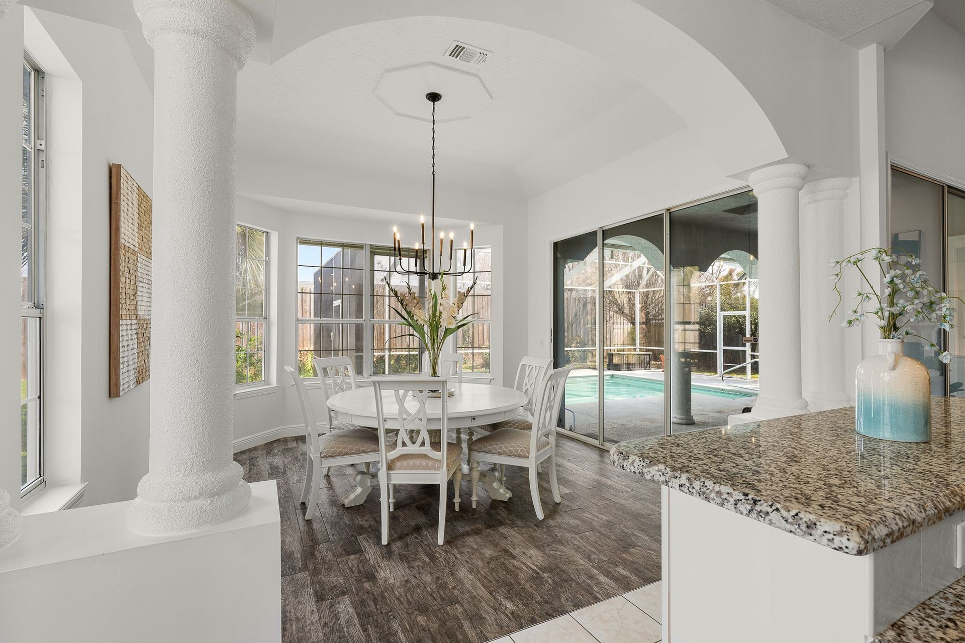 Bright dining room with white chairs, arched columns, granite counter, and a pool visible through open doors