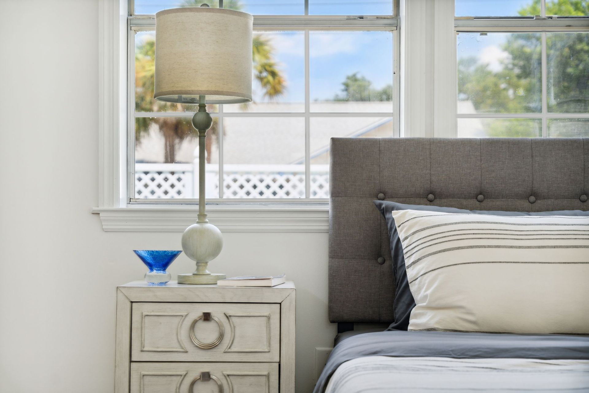Bright bedroom with gray tufted bed, white nightstand, lamp, and window view