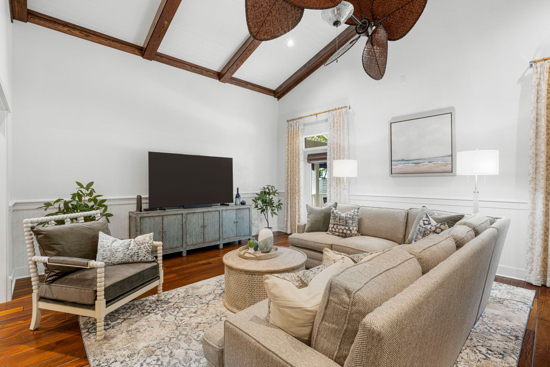Bright living room with beige sofas, TV, woven chair, and high wood-beamed ceiling