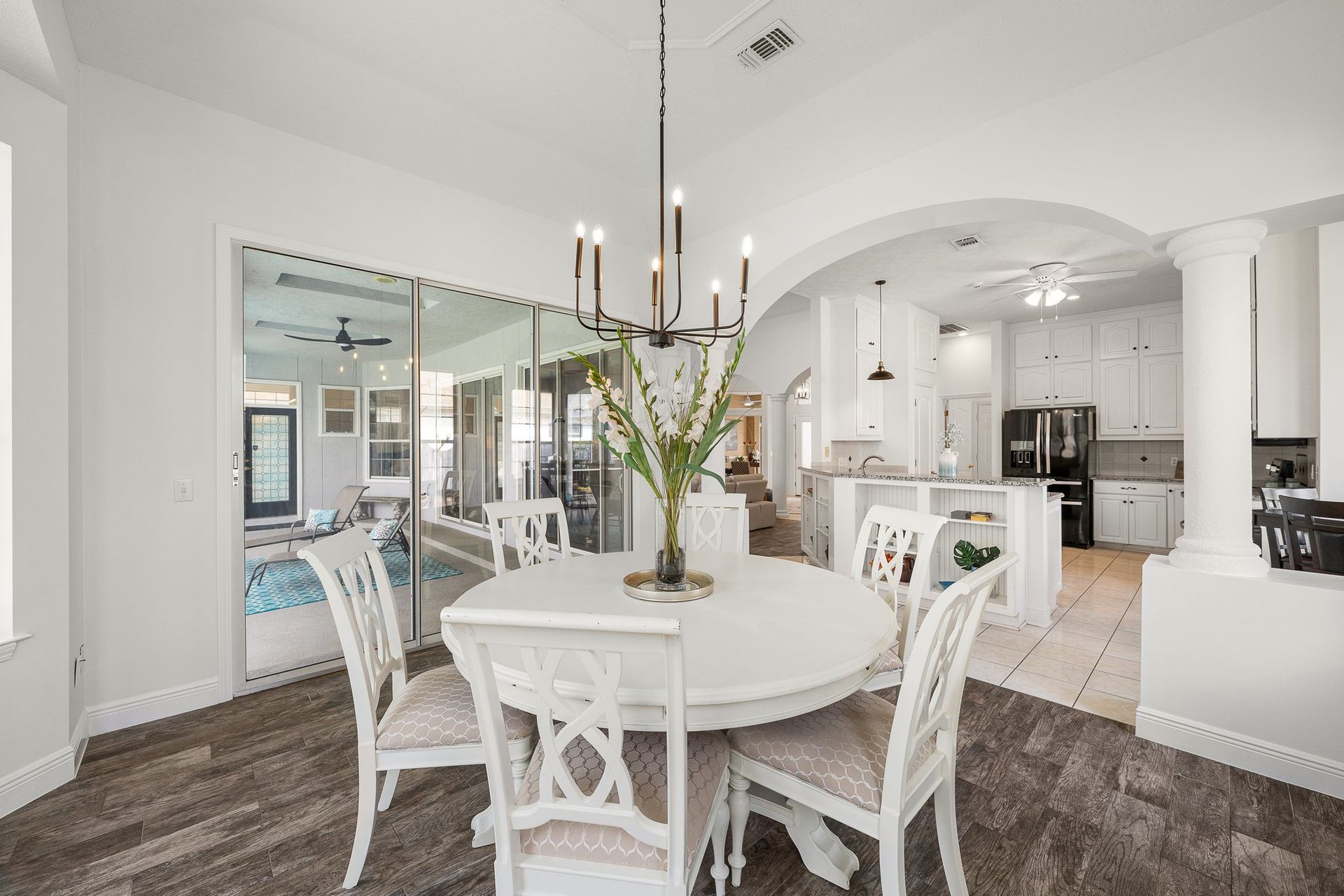 Bright open dining room with white chairs, round table, chandelier, and kitchen beyond