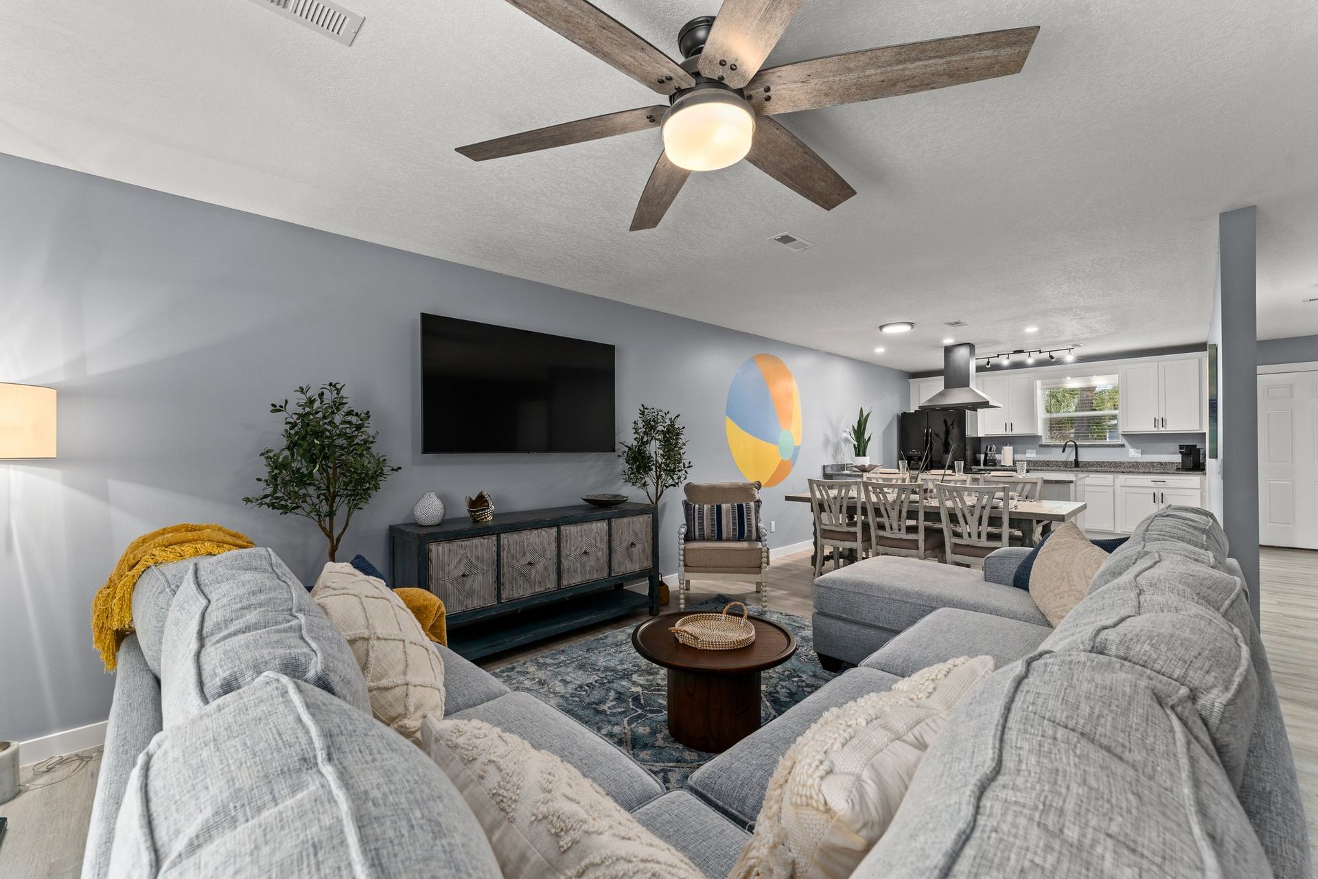 Bright living room with gray sofas, wall-mounted TV, ceiling fan, and open kitchen beyond