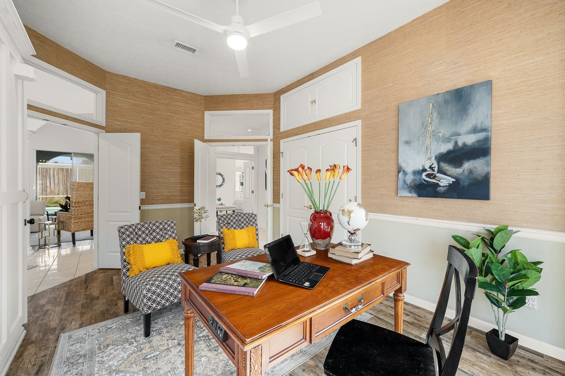 Cozy home office with a wooden desk, patterned chairs, wall art, and potted plants in a bright room