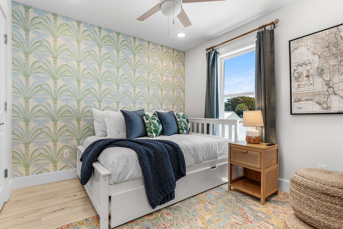 Bright bedroom with patterned wallpaper, twin bed, wicker nightstand, window, and colorful rug