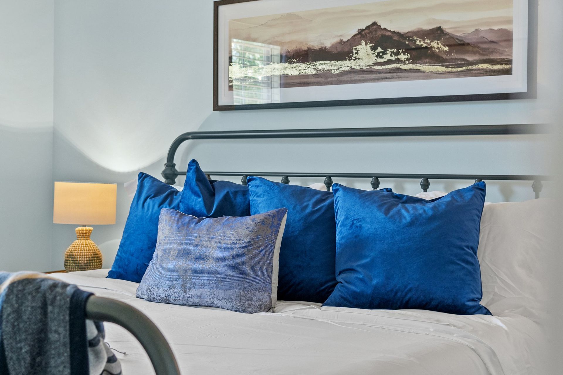 Bedroom with a white bed, blue pillows, lamp, and framed landscape art above the headboard.