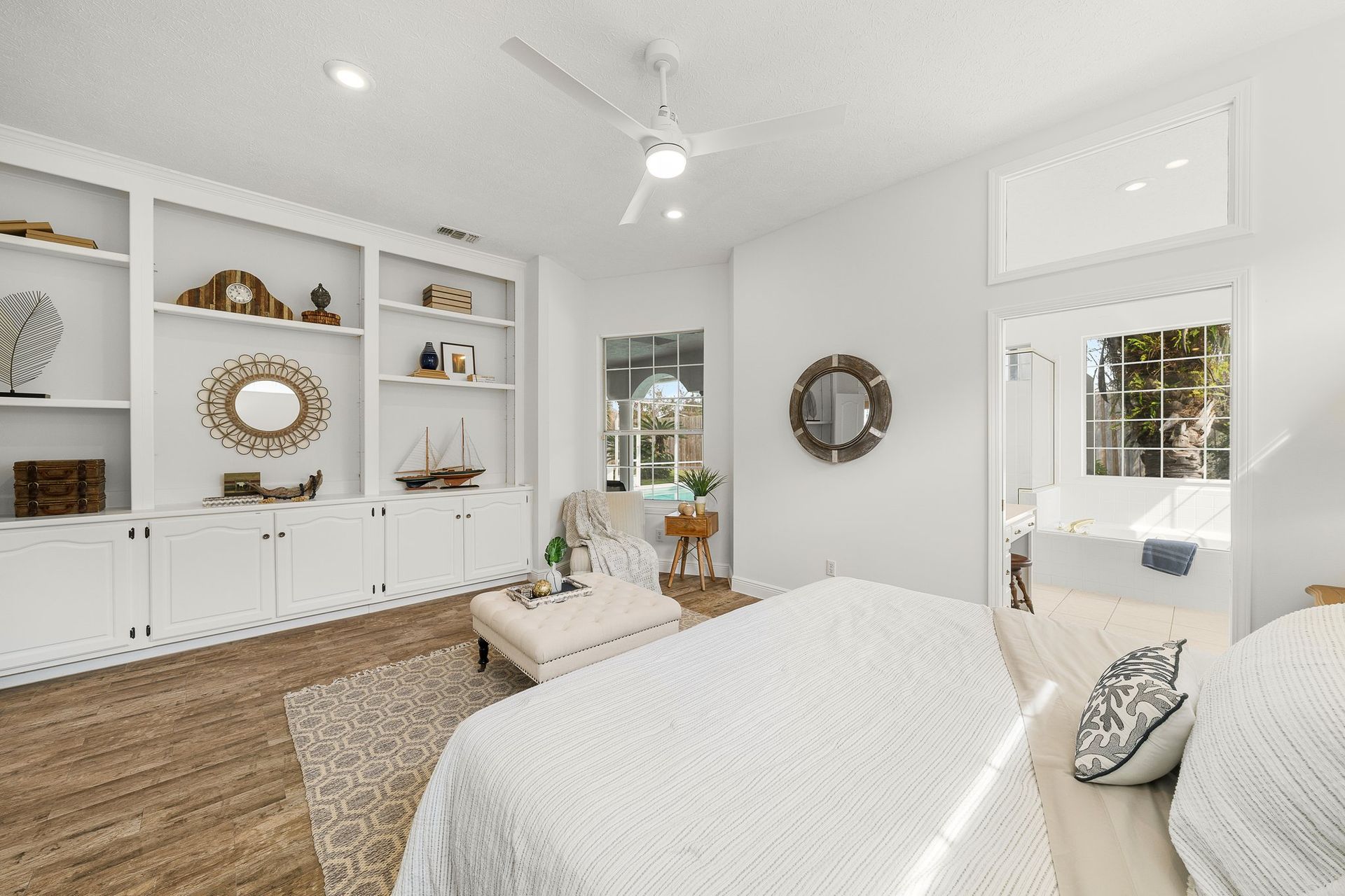 Bright white bedroom with built-in shelves, woven rug, round mirrors, and a bed by a window.