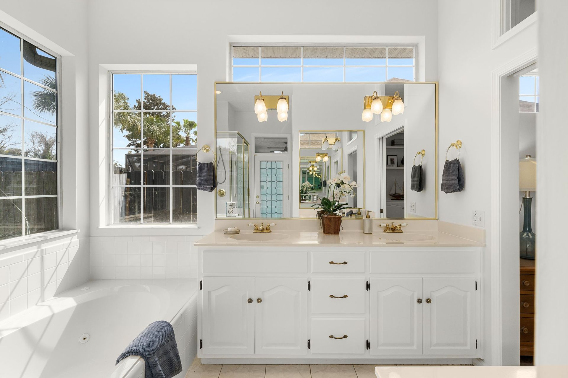 Bright white bathroom with double vanity, large mirrors, windows, and brass light fixtures