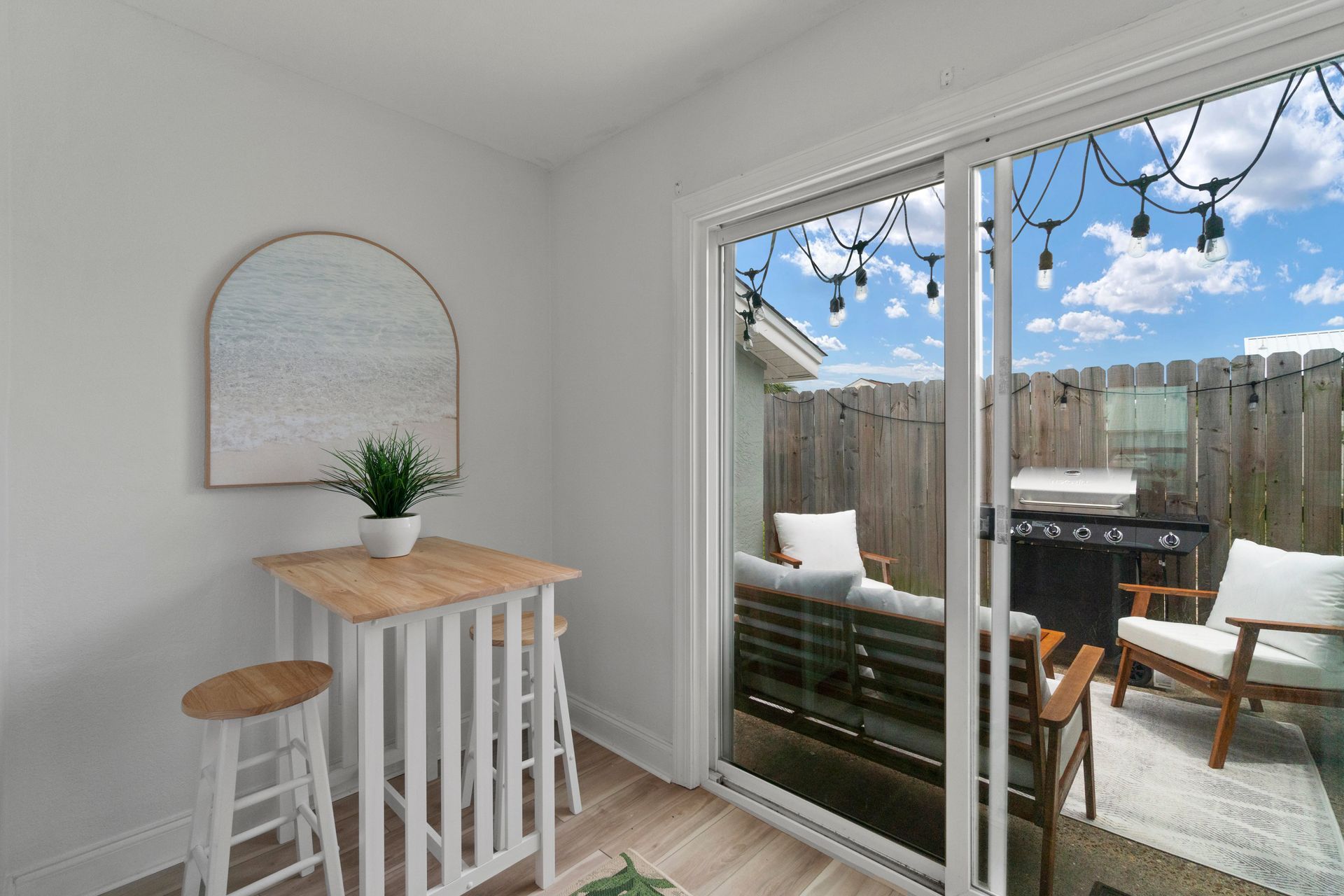 Small bright nook with a wood table, stool, round mirror, and sliding door to a fenced patio with chairs.