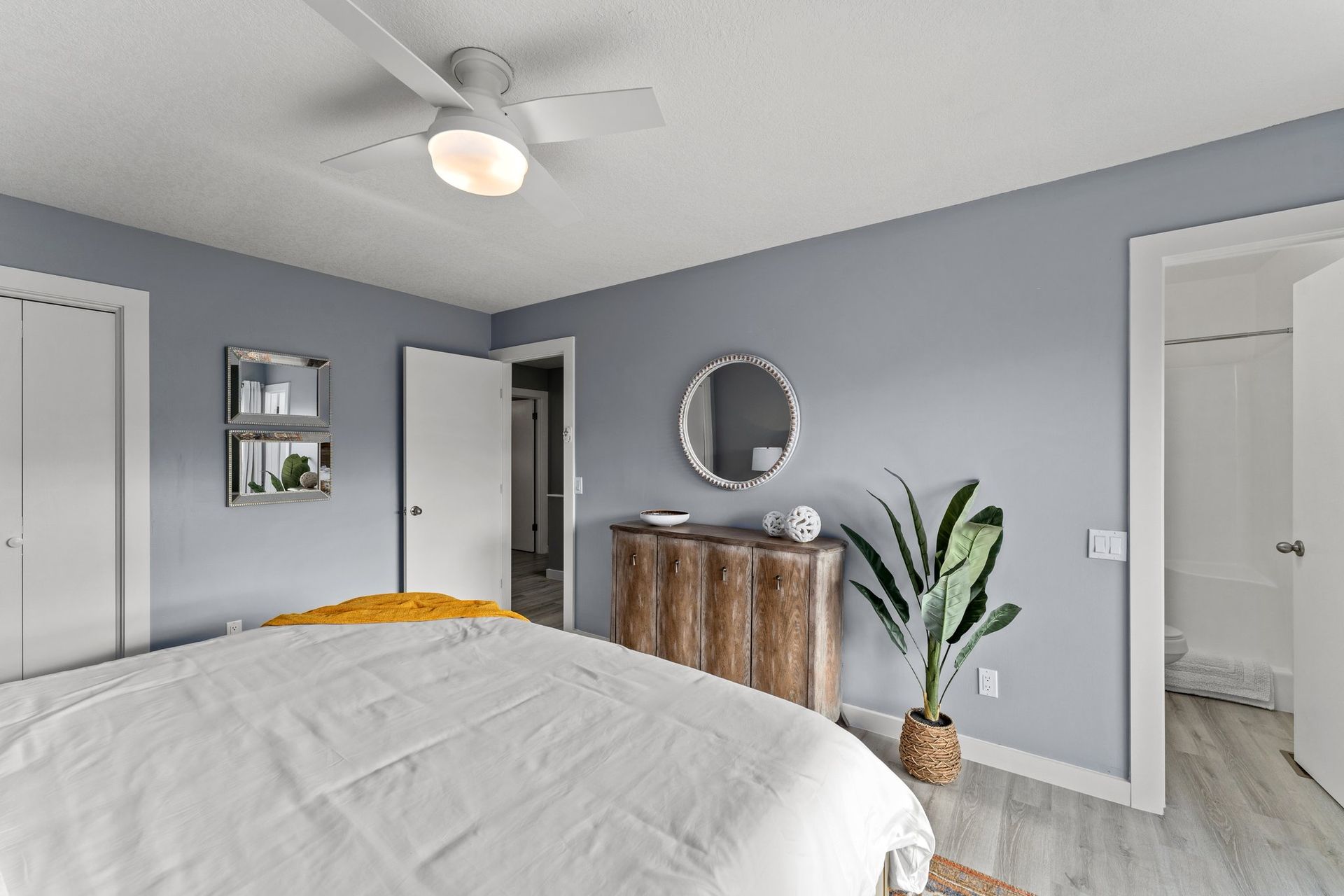 Bright bedroom with gray walls, white bed, wooden dresser, round mirror, and potted plant.