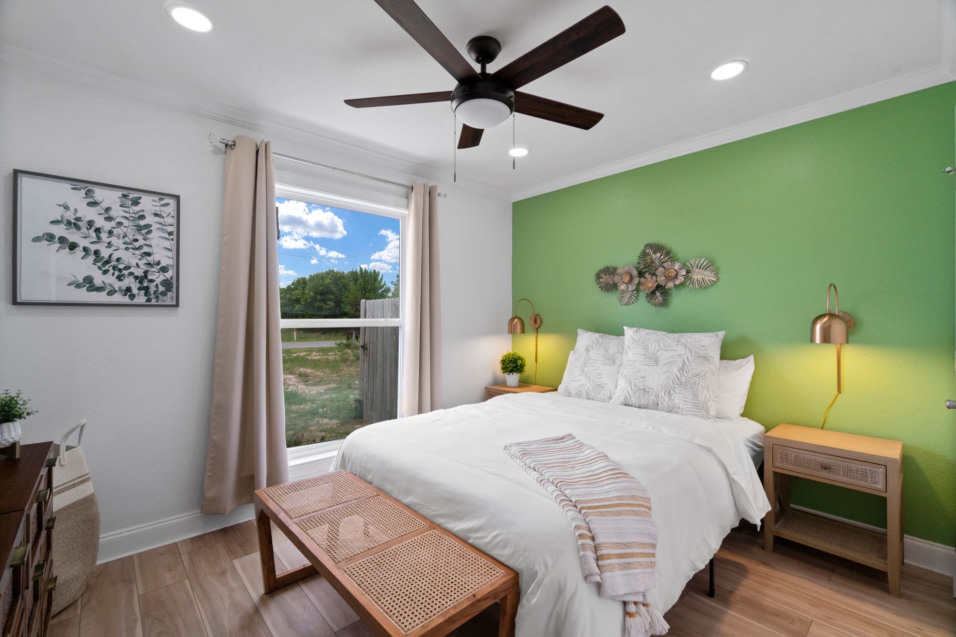 Bright bedroom with white bed, green accent wall, wood furniture, and a window view of trees and clouds.