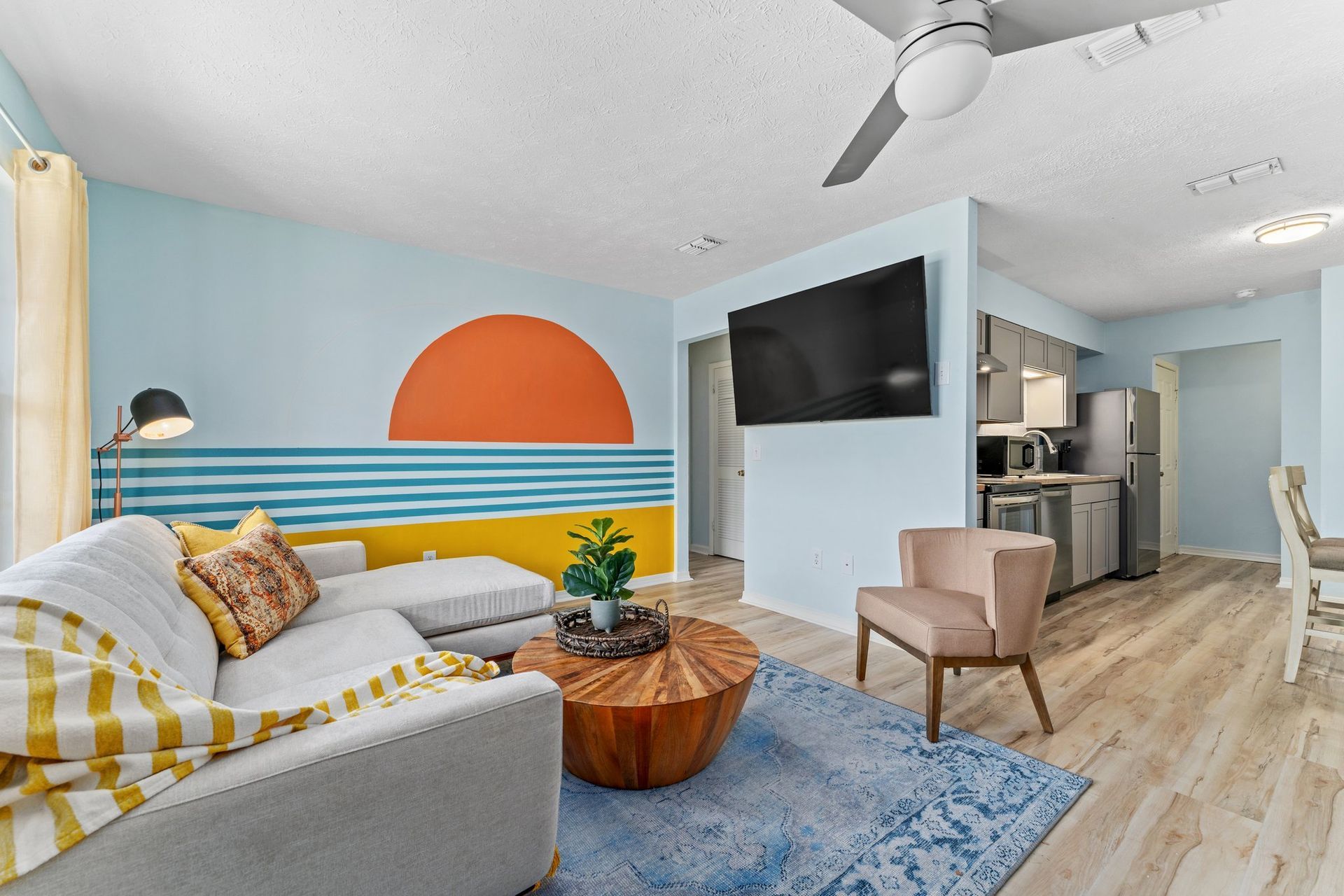 Bright living room with striped sofa, round wood coffee table, blue rug, wall TV, and kitchenette.