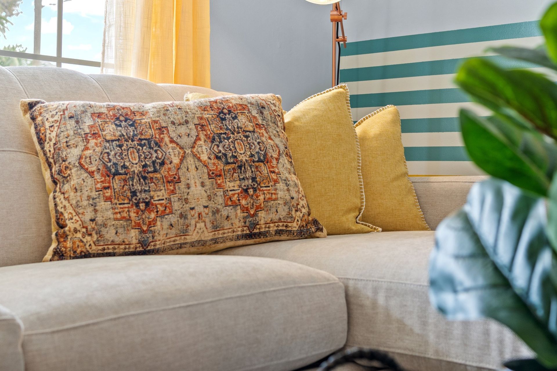 Beige sofa with patterned and yellow pillows beside a large green plant in a bright living room