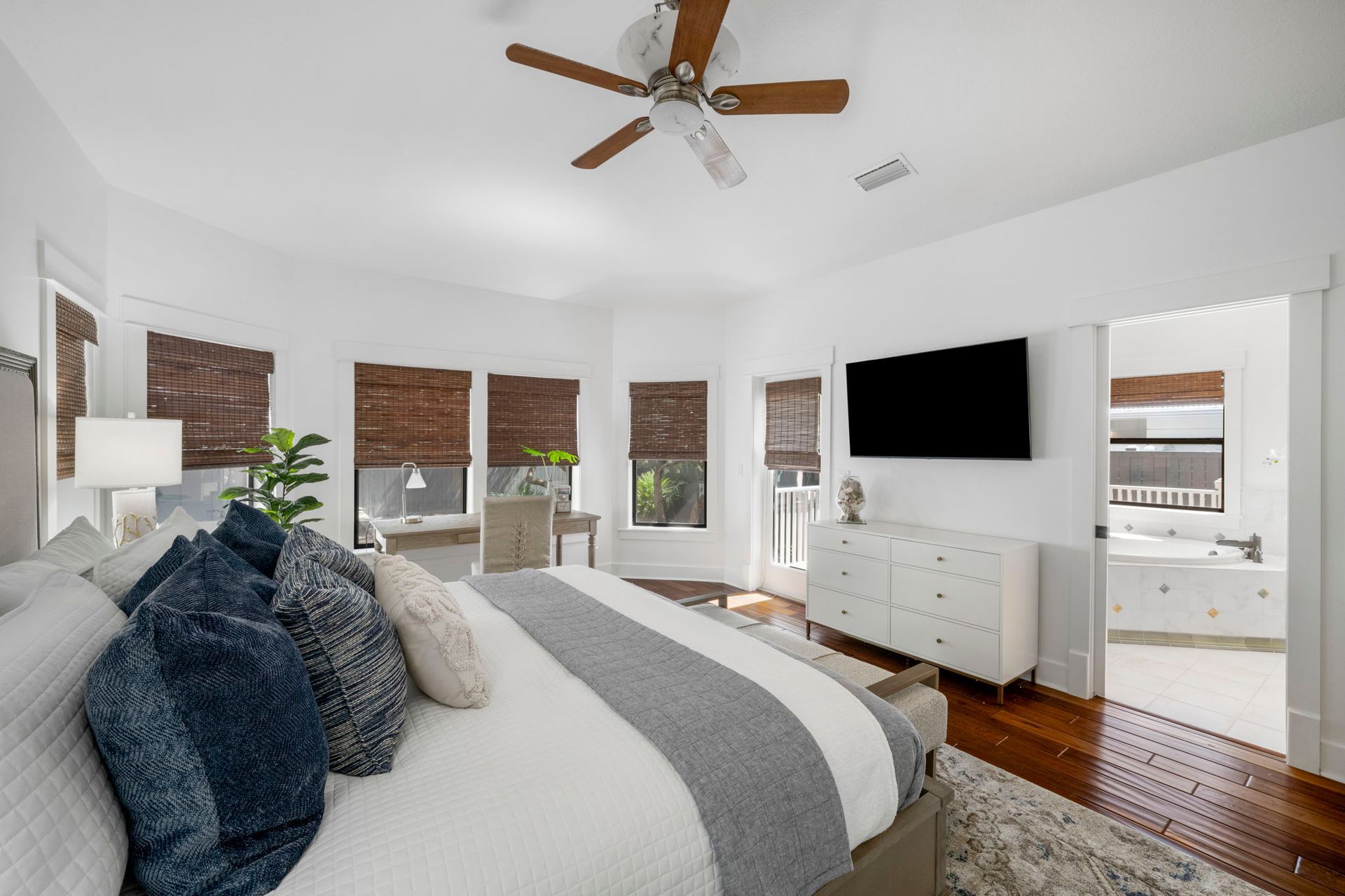 Bright bedroom with gray bed, white dresser, wall-mounted TV, ceiling fan, and large windows with blinds