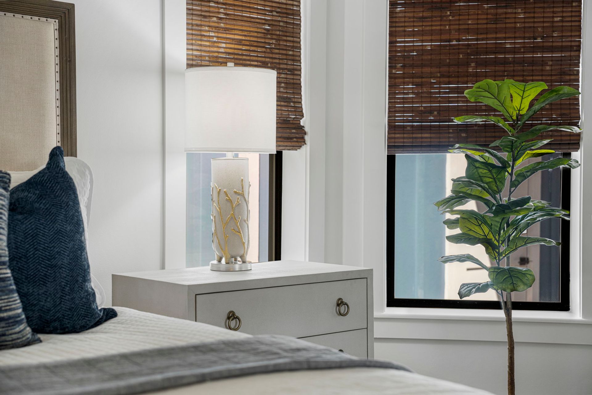 Modern bedroom corner with bed, bedside lamp, dresser, and tall potted plant by the window.