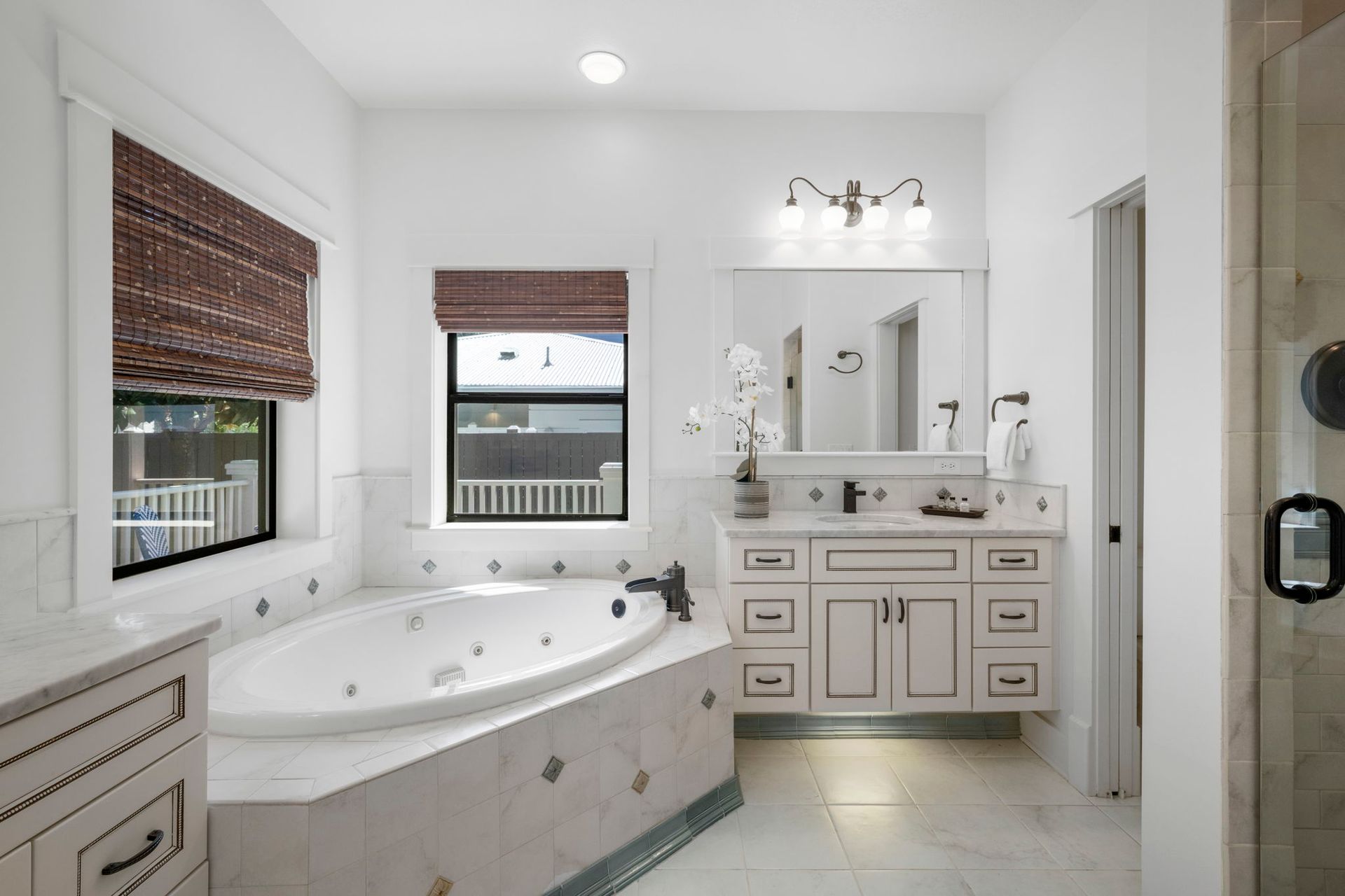 Bright white bathroom with a corner soaking tub, vanity, mirror, windows, and woven blinds.