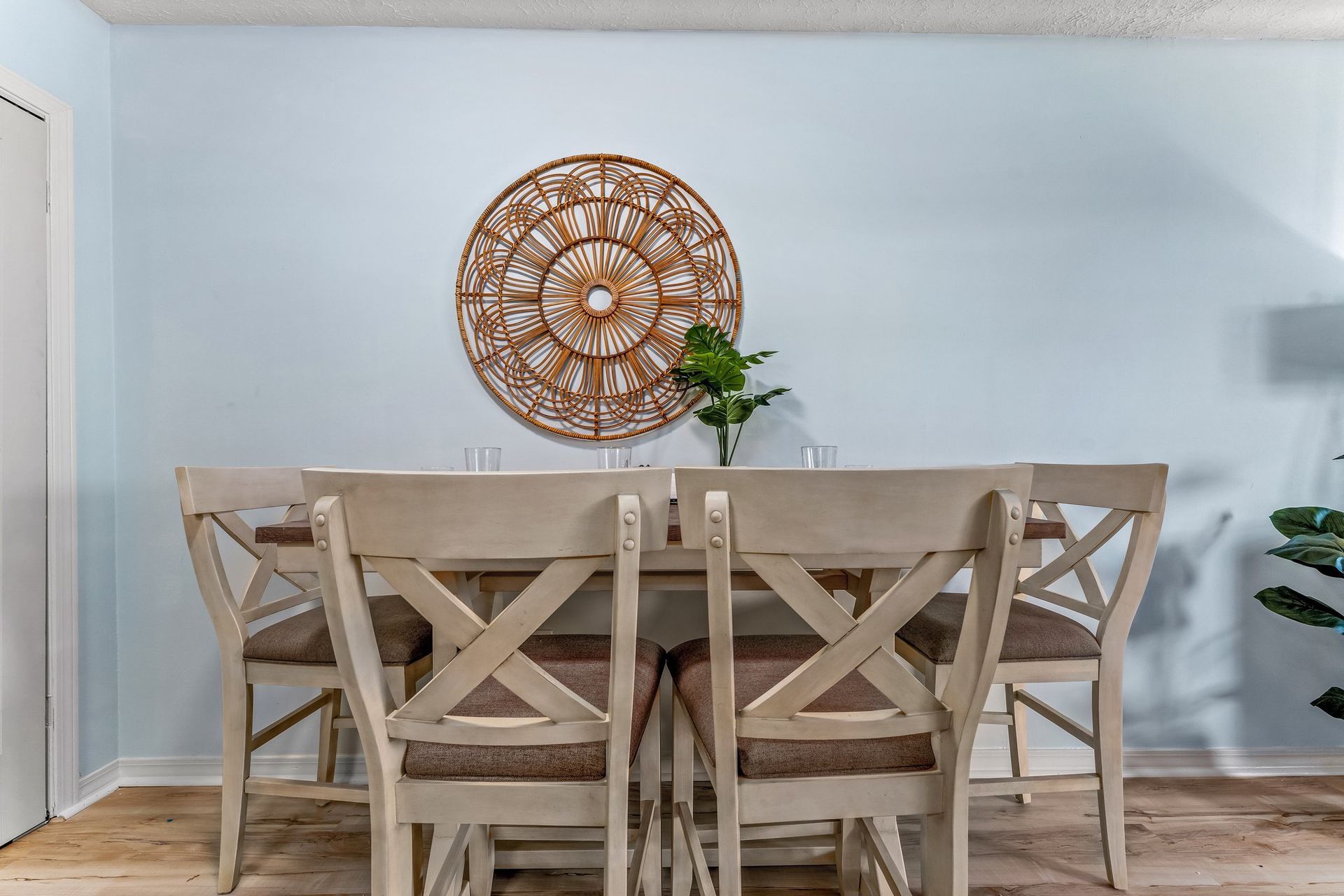Dining room with rustic wooden table and chairs against a light blue wall, centered by a woven wall decor and plant.