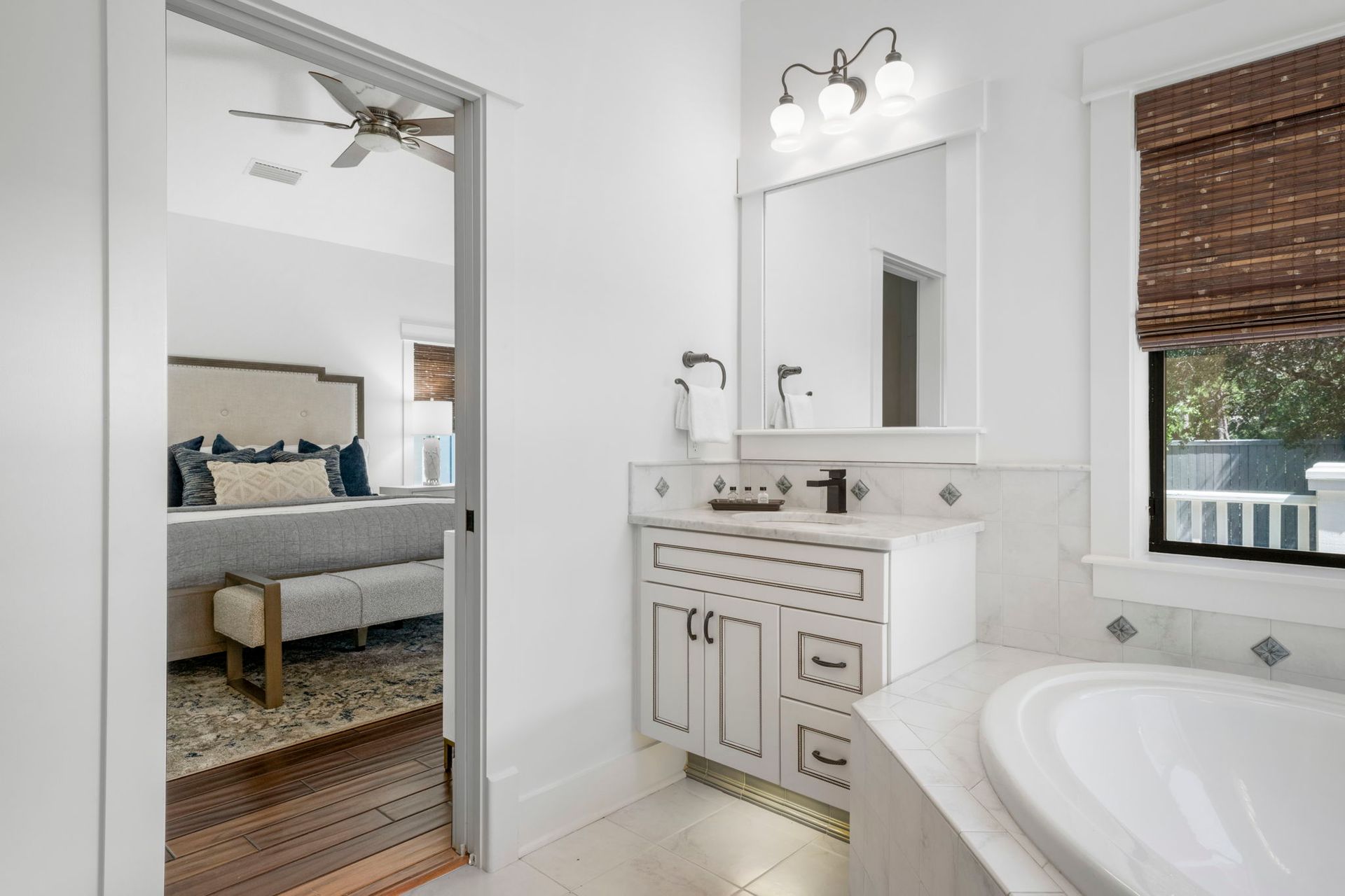 Bright bathroom with white vanity, mirror, tub, and a doorway to a bedroom with gray bed and rug