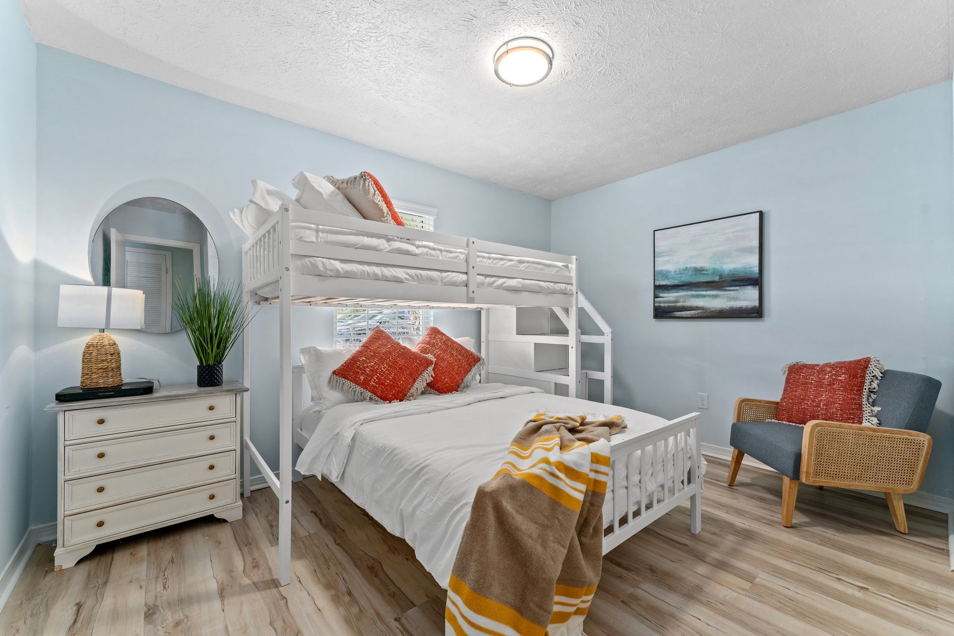 Bright bedroom with white bunk bed, blue walls, wicker chair, dresser, and coastal decor.