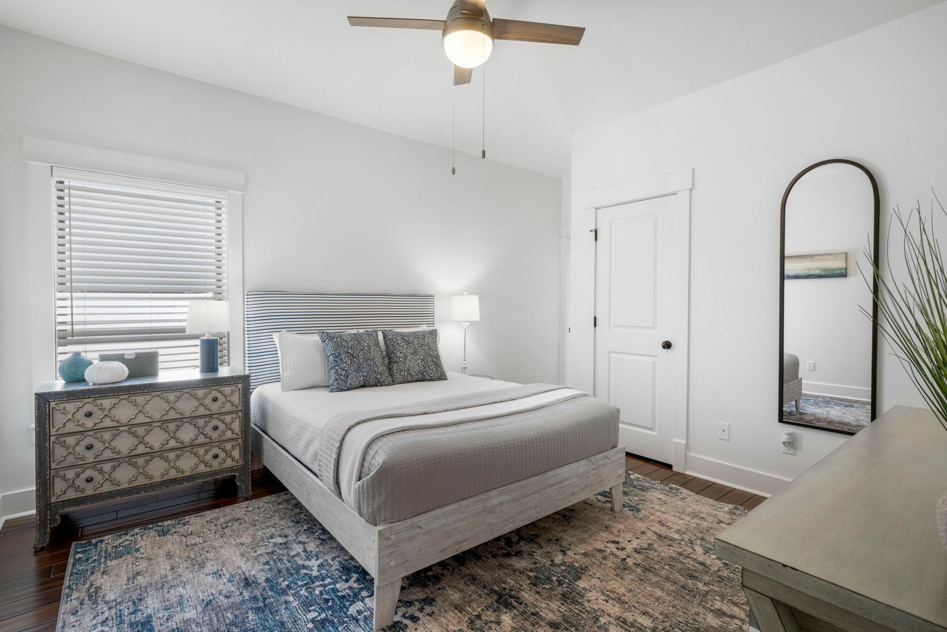 Bright bedroom with gray bed, dresser, mirror, window blinds, and blue rug