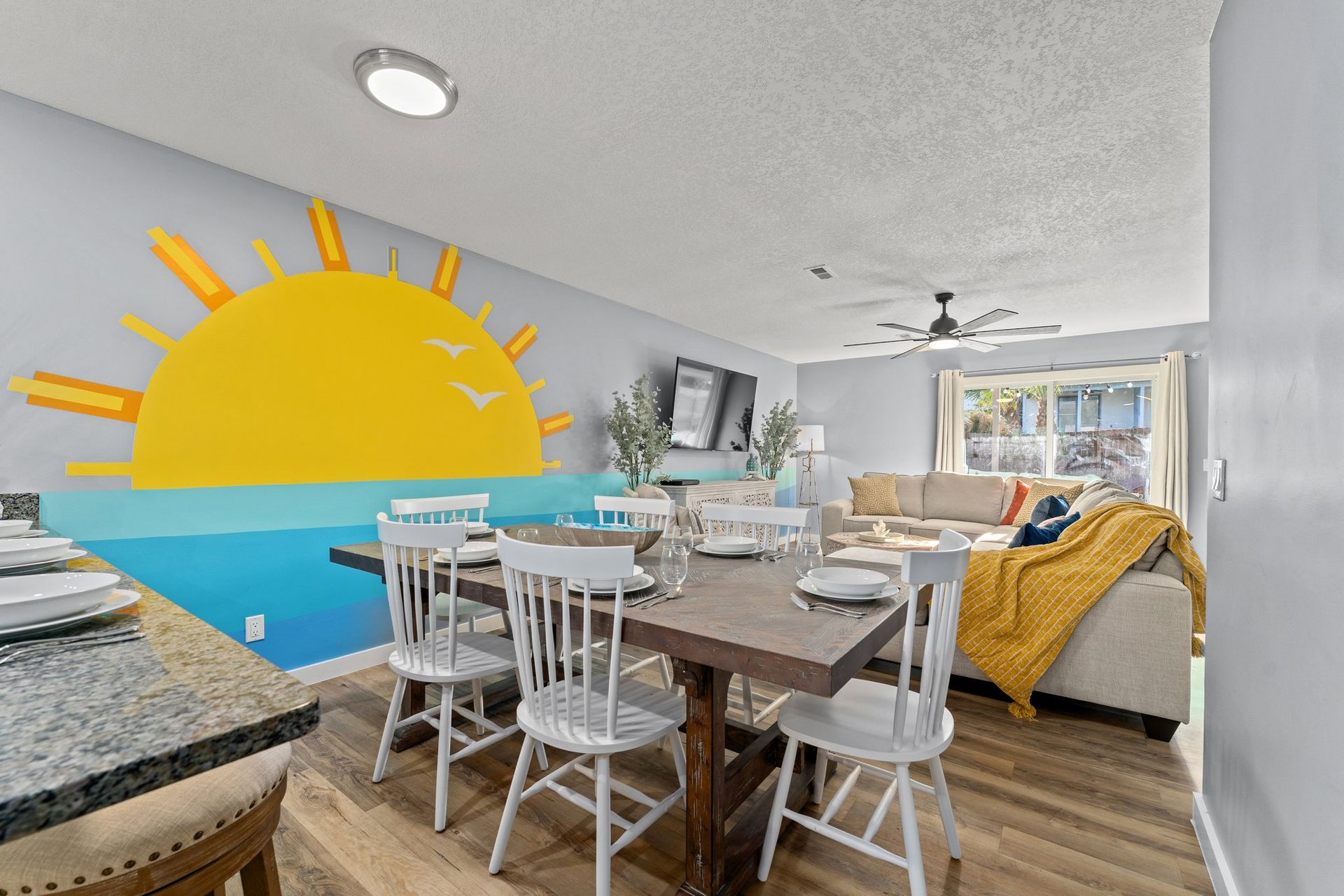 Bright dining room with beach mural, white chairs, wood table, and a mustard-yellow sofa.
