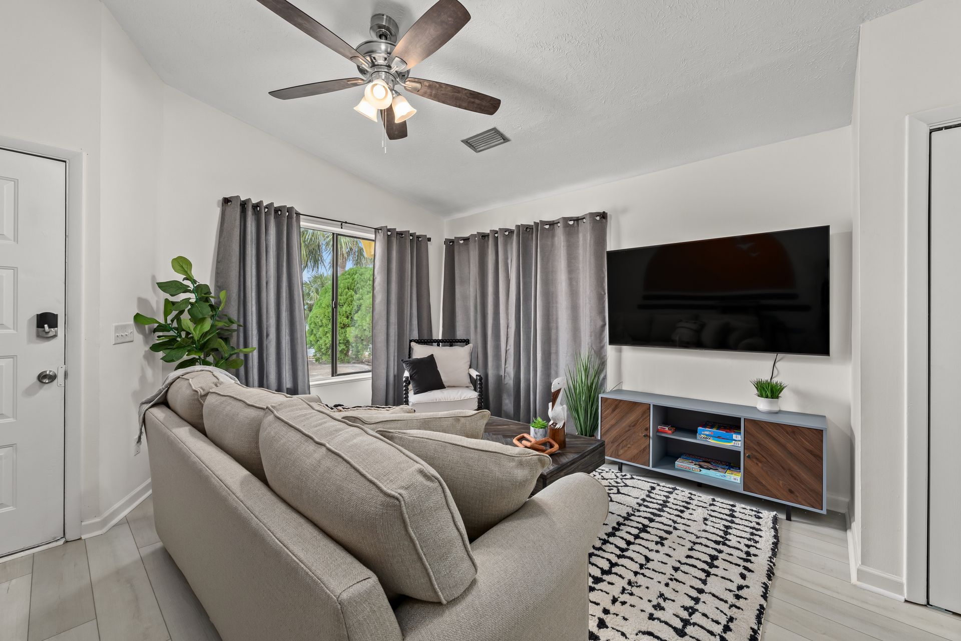 Bright living room with gray sectional, wall-mounted TV, ceiling fan, and sliding glass doors with curtains