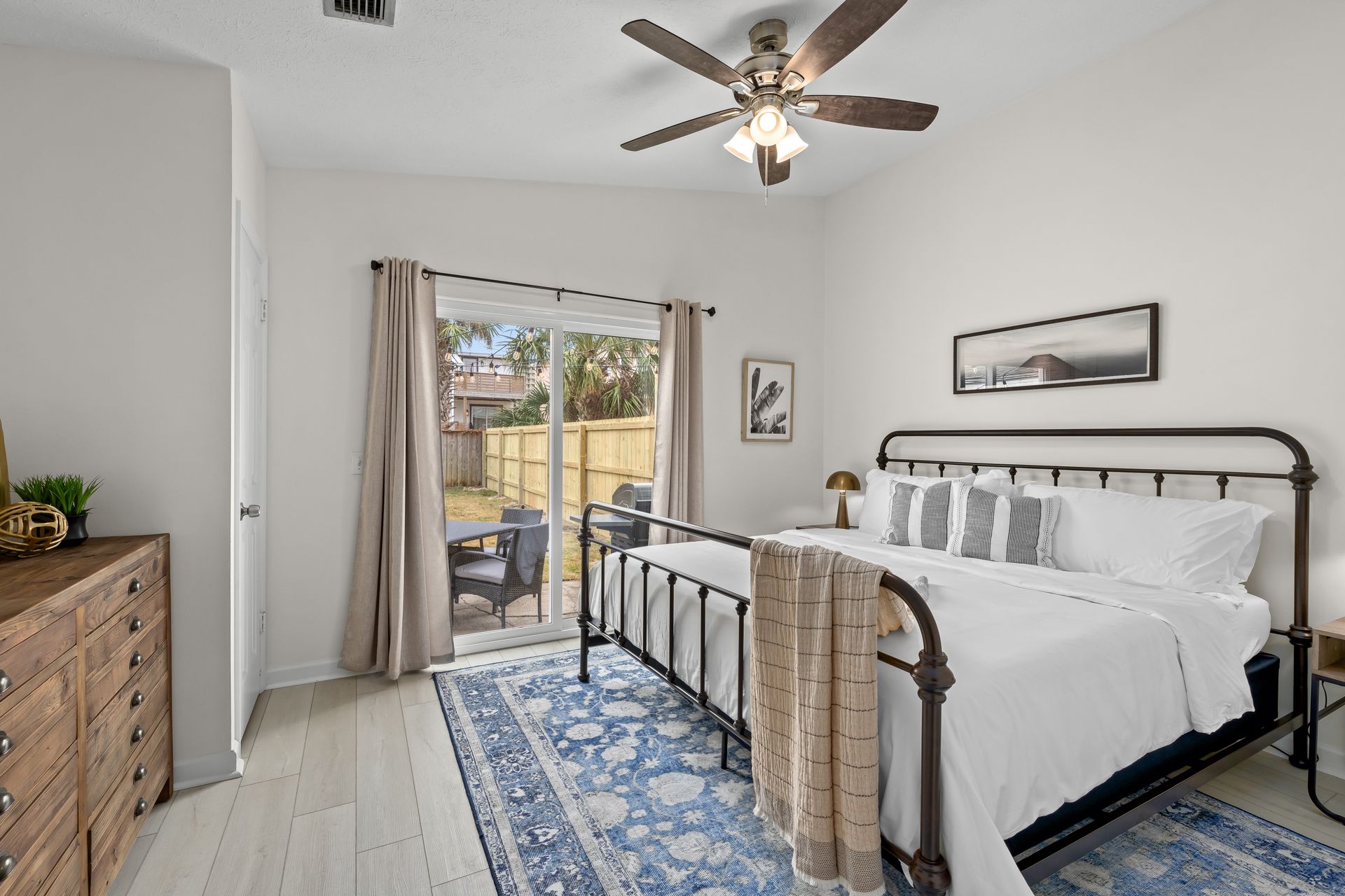 Bright bedroom with a black metal bed, blue rug, dresser, ceiling fan, and balcony doors