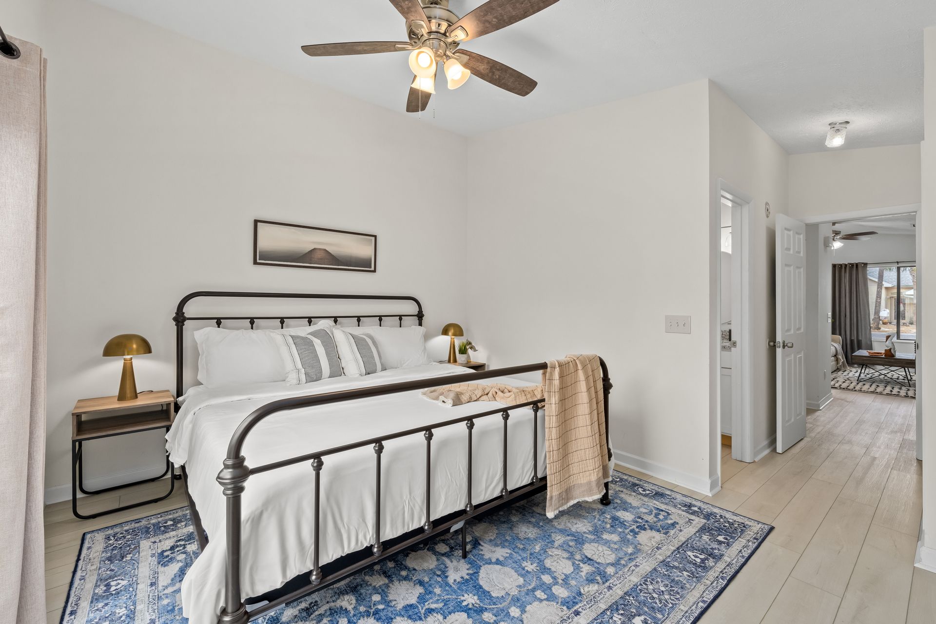 Bright bedroom with a black metal bed, white bedding, blue rug, ceiling fan, and hallway view