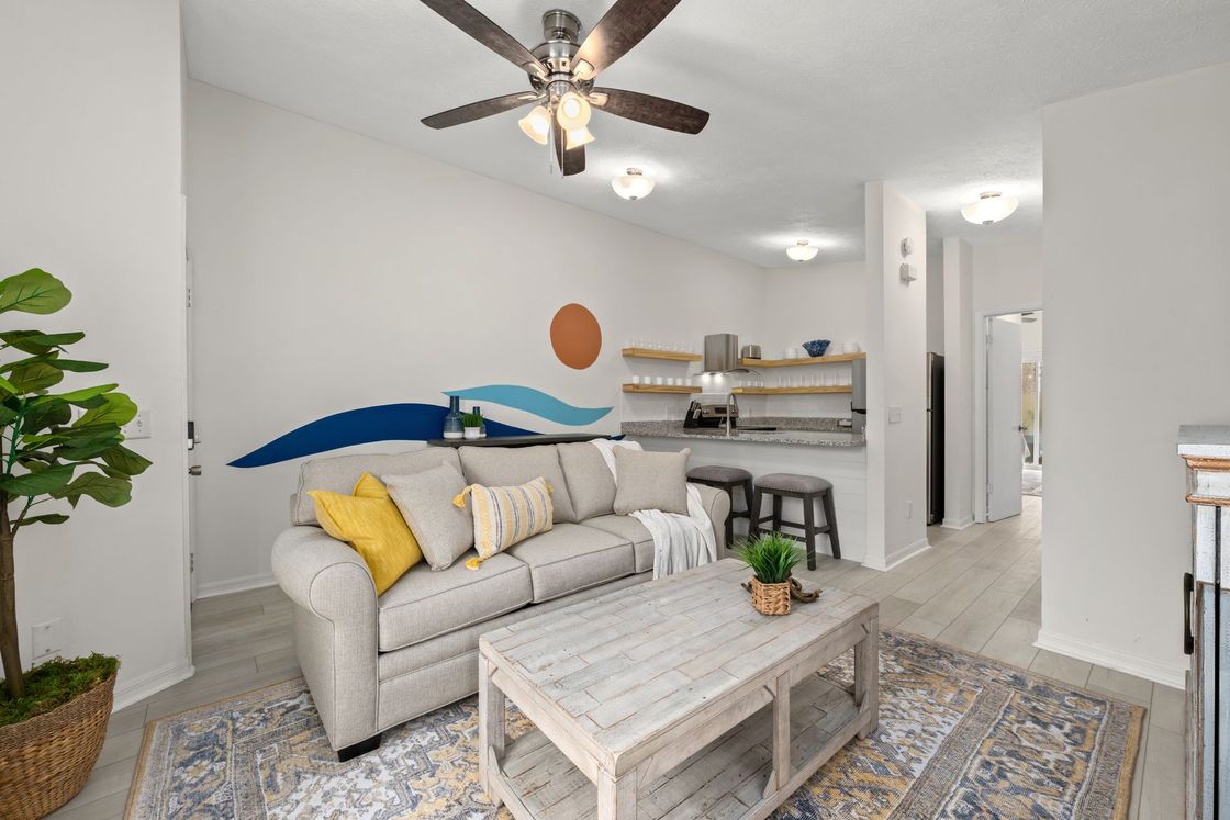 Bright living room with gray sofa, coffee table, patterned rug, wall art, ceiling fan, and small kitchen area