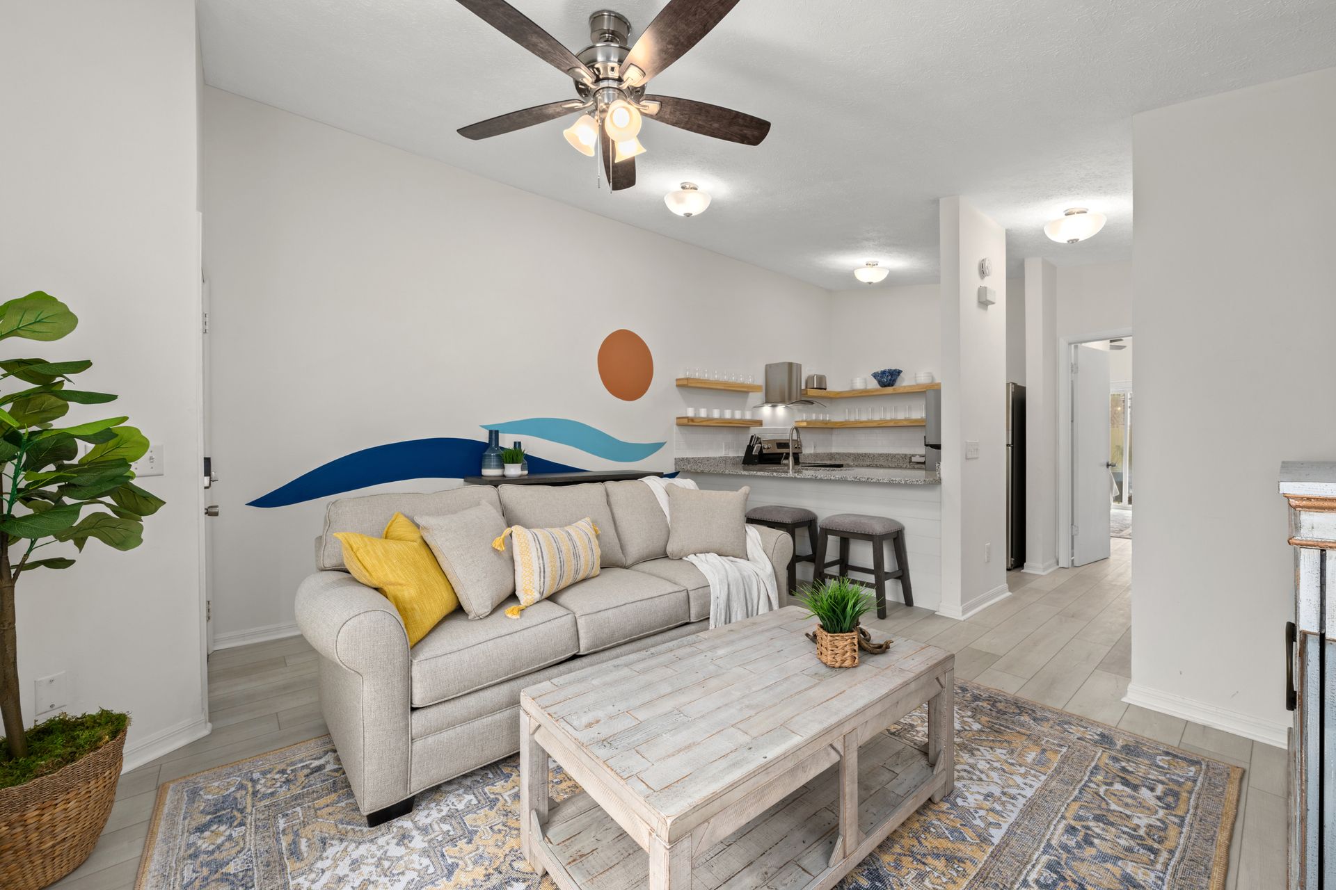 Bright living room with gray sofa, coffee table, patterned rug, wall art, ceiling fan, and small kitchen area