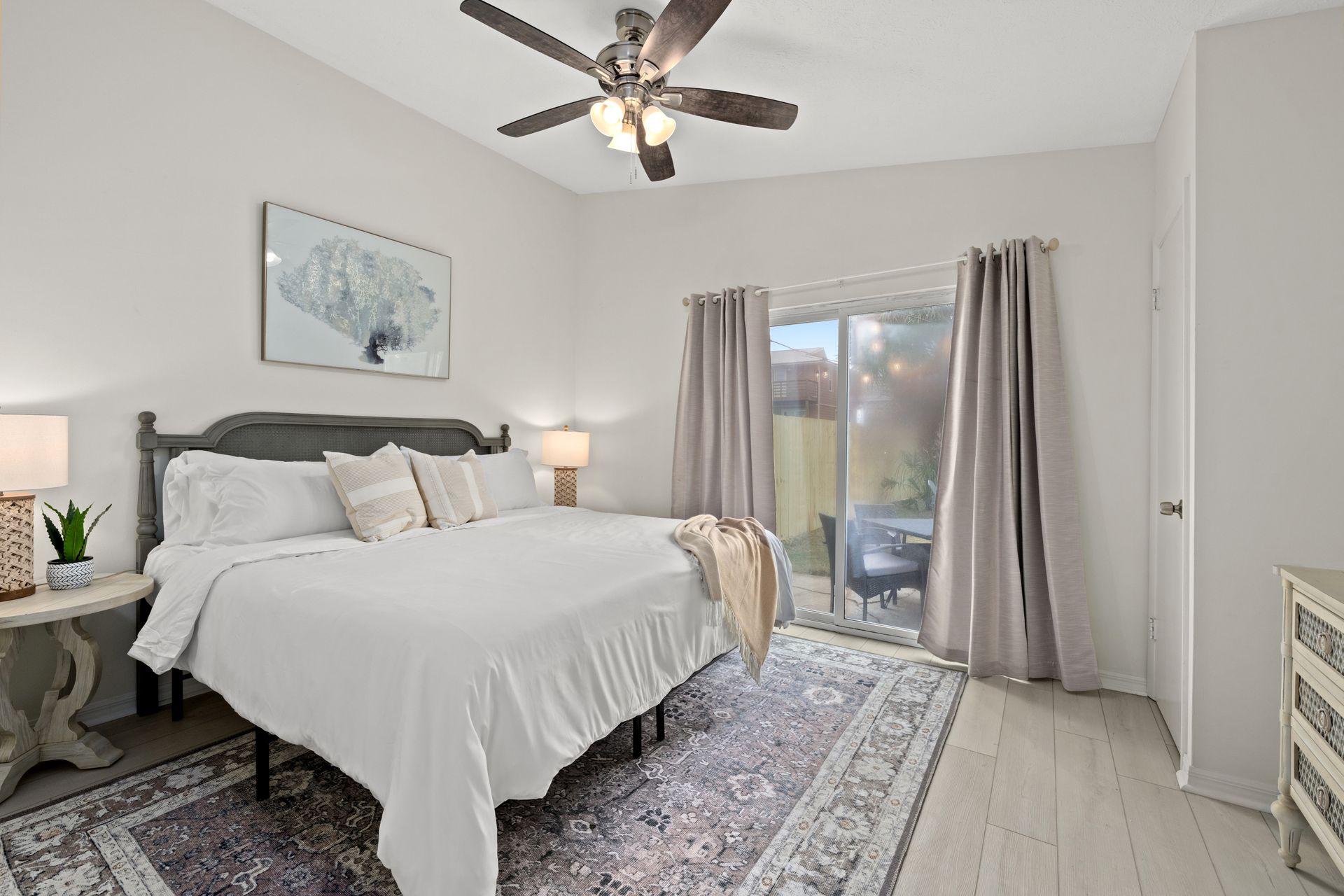 Bright bedroom with a made bed, gray curtains, ceiling fan, and sliding glass door.