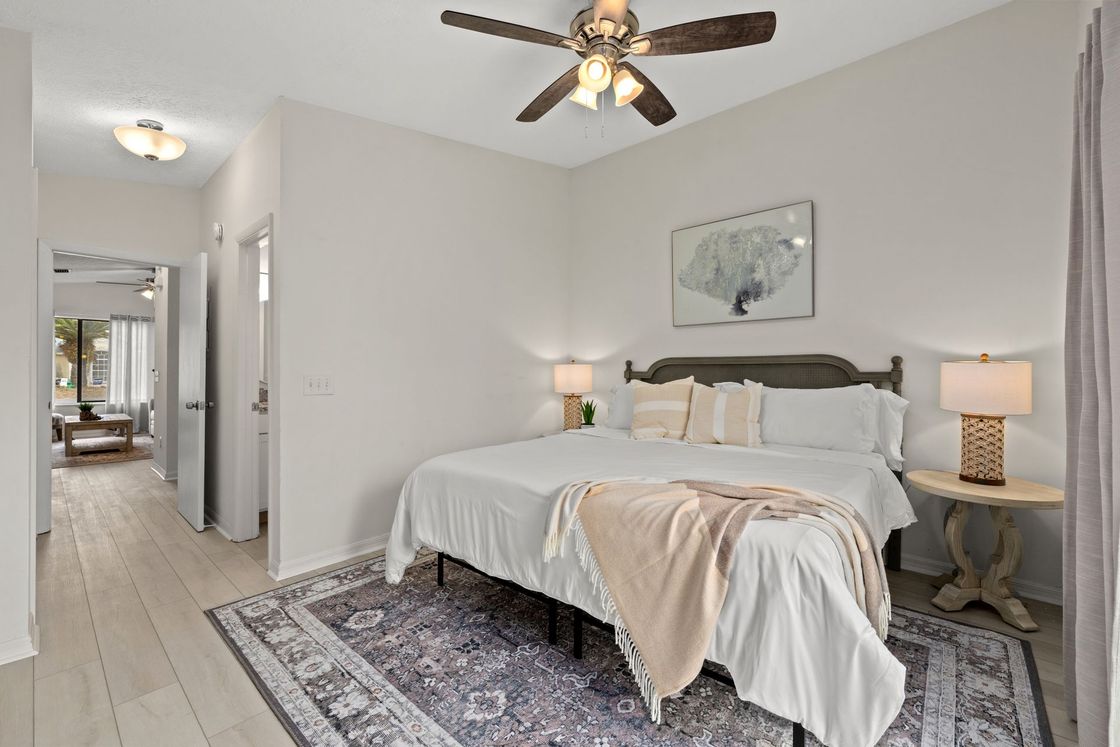 Bright bedroom with a bed, bedside lamps, ceiling fan, rug, and hallway view