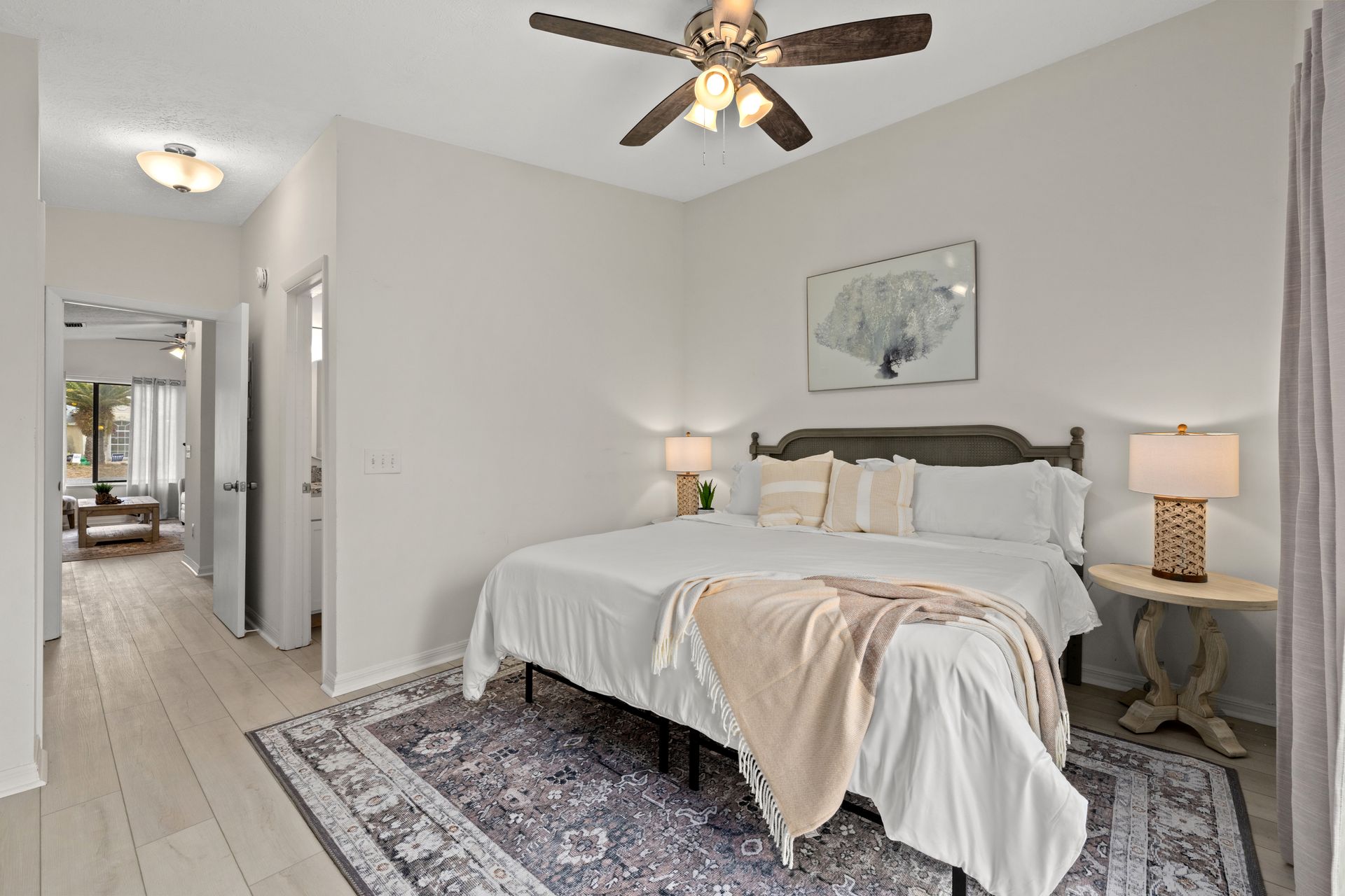 Bright bedroom with a bed, bedside lamps, ceiling fan, rug, and hallway view