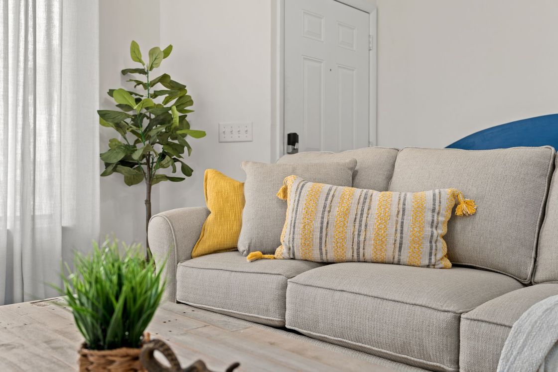 Bright living room with a beige sofa, yellow pillows, a striped cushion, and a potted plant by the window.