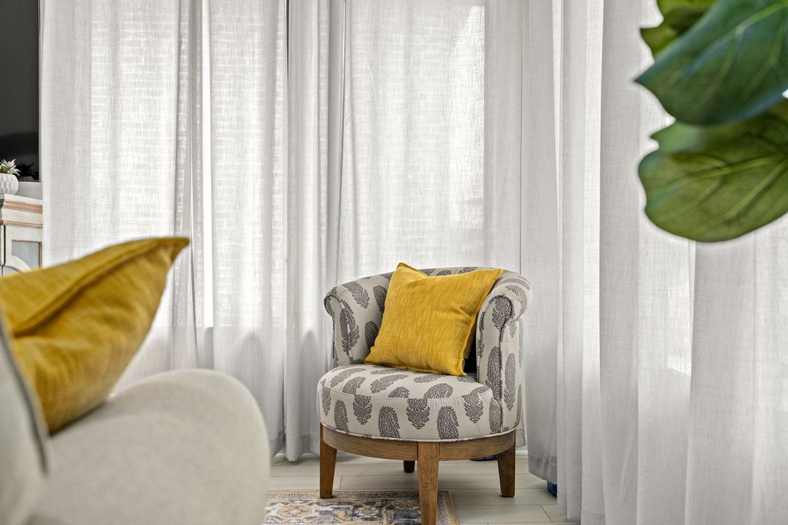 Cozy living room chair with yellow pillow beside sheer white curtains and a patterned rug