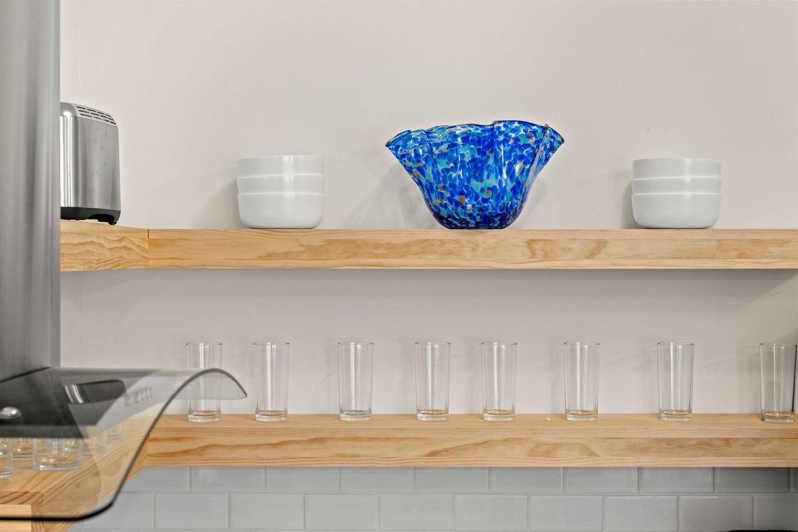 Kitchen shelf with clear glasses and white bowls, centered by a blue glass bowl on a wooden shelf.