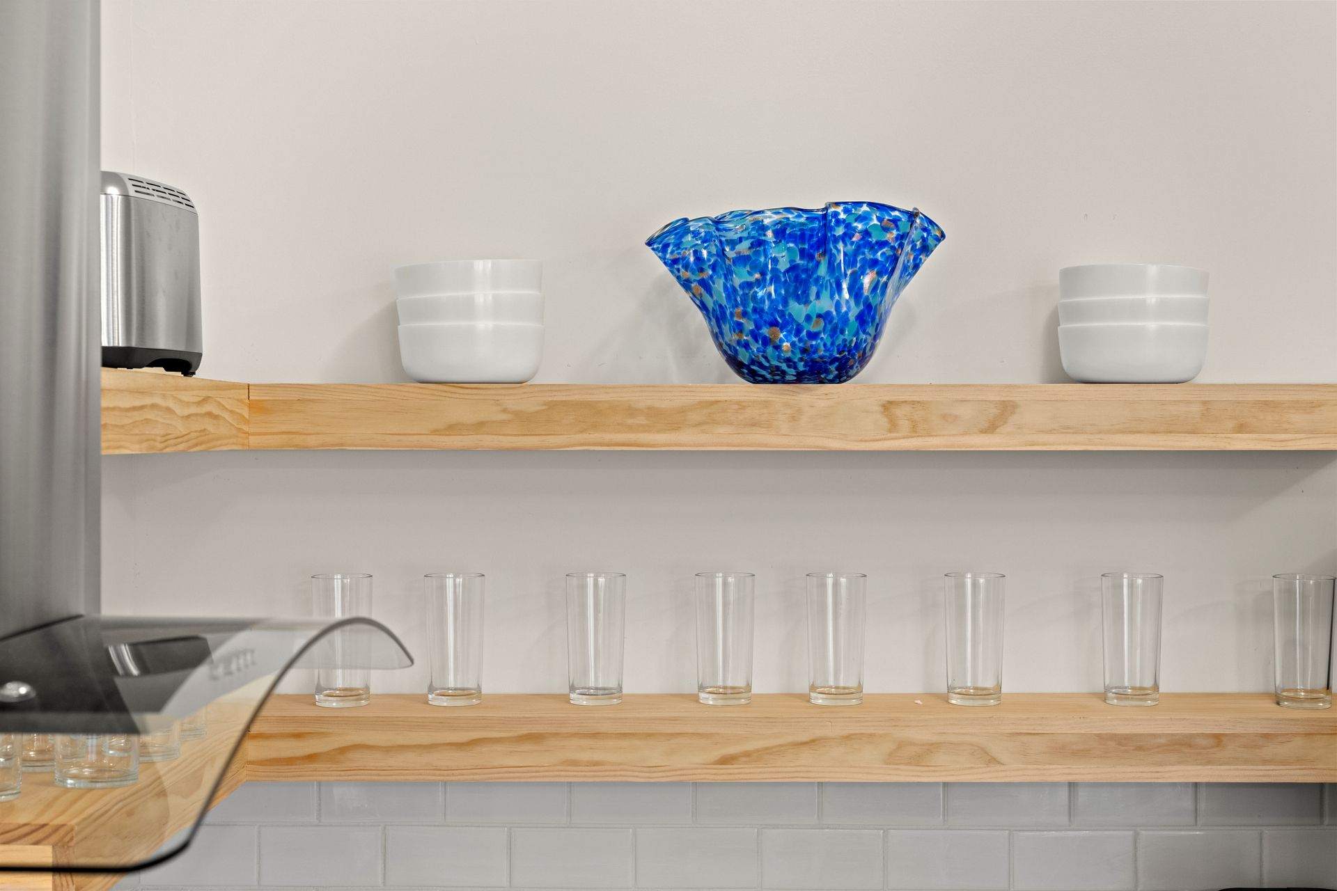 Kitchen shelf with clear glasses and white bowls, centered by a blue glass bowl on a wooden shelf.