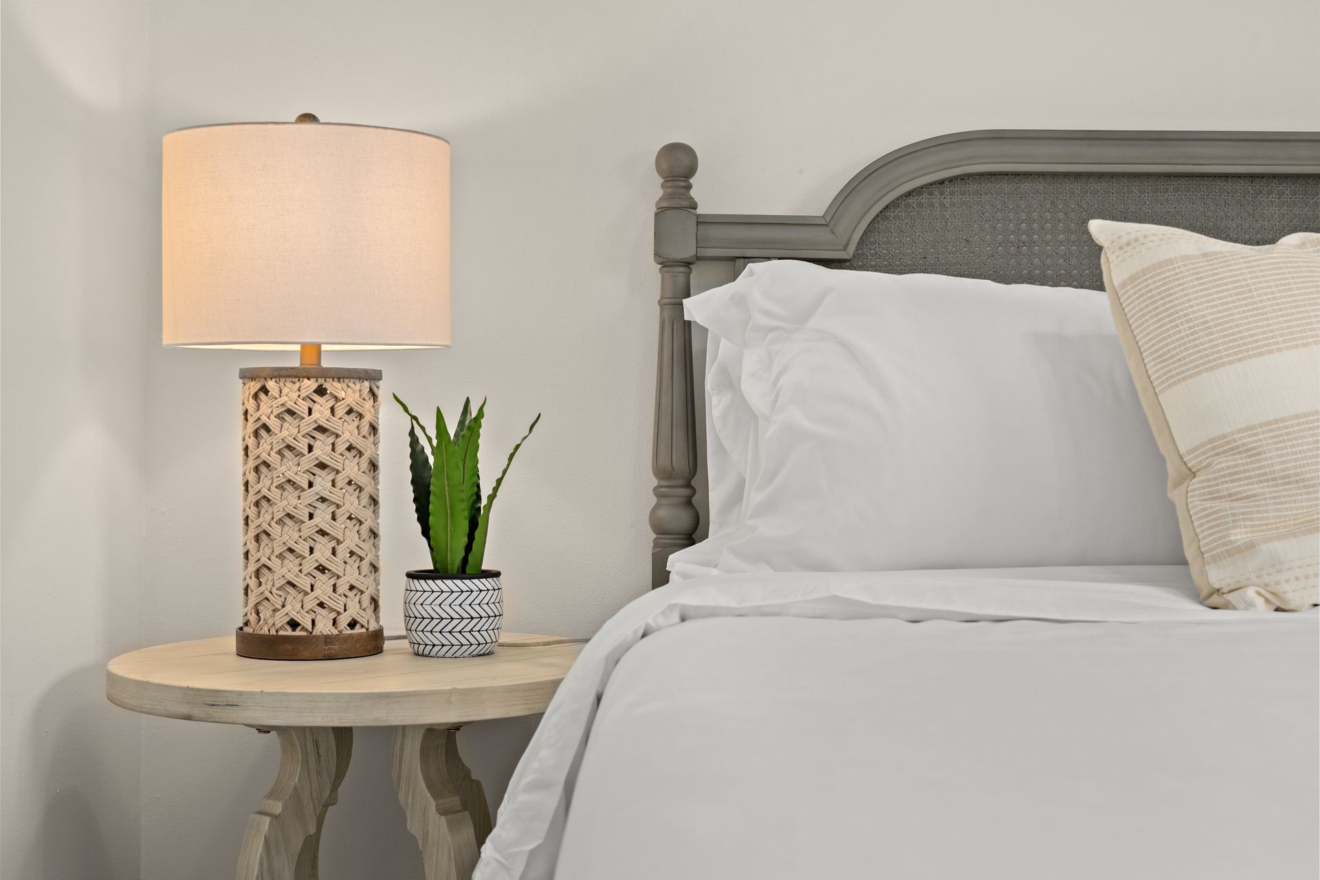 Neatly made bed beside a bedside table with lamp and small potted plant in a soft gray bedroom