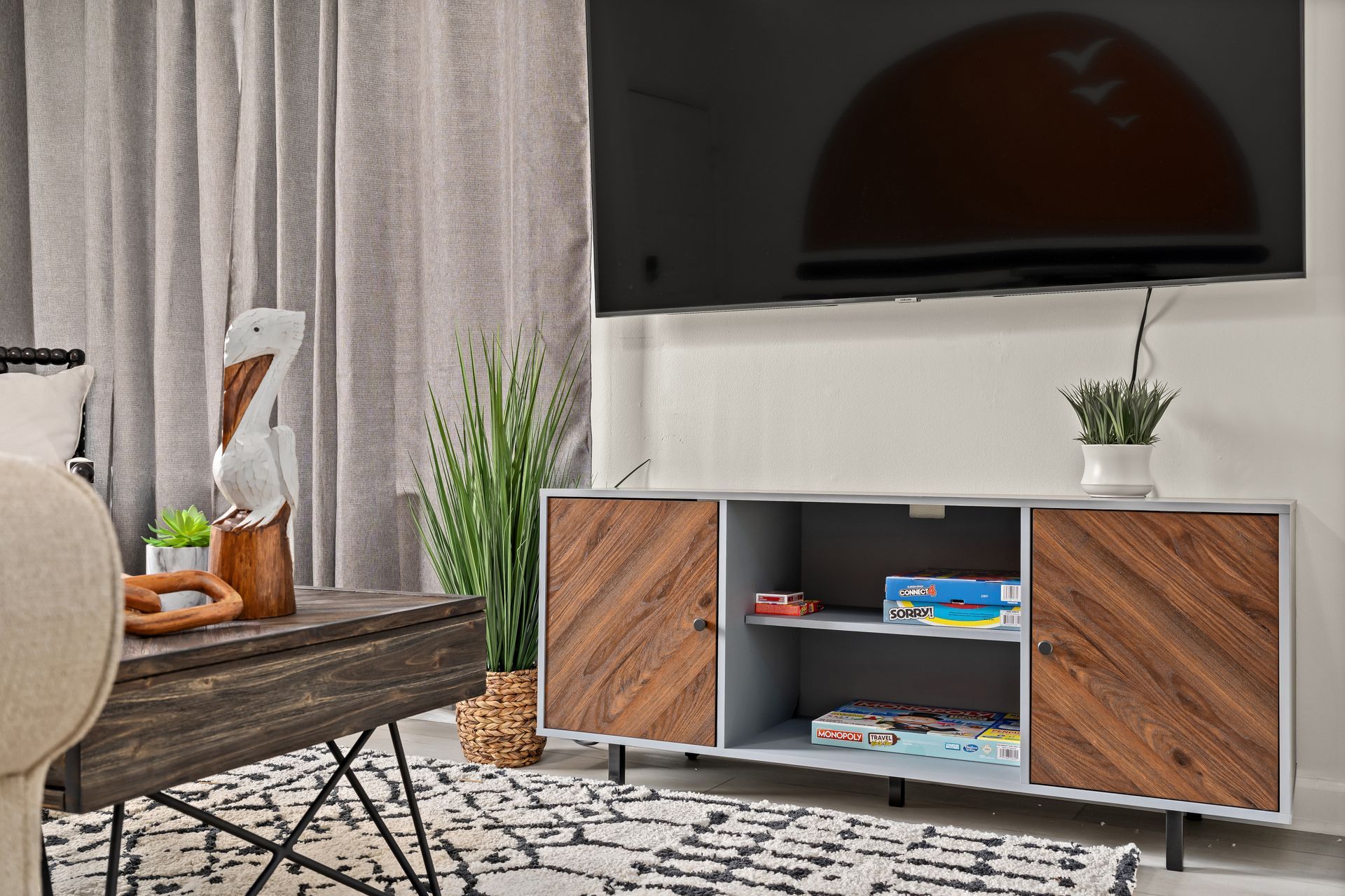 Modern living room with wall-mounted TV, wood media console, curtains, rug, plant, and chair