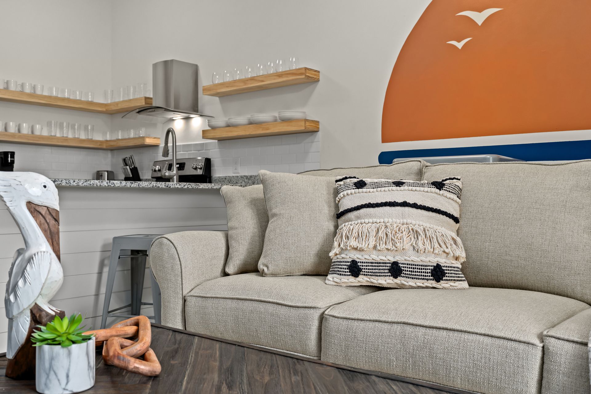 Modern living room with gray sofa, patterned pillows, orange accent wall, and open kitchen shelves