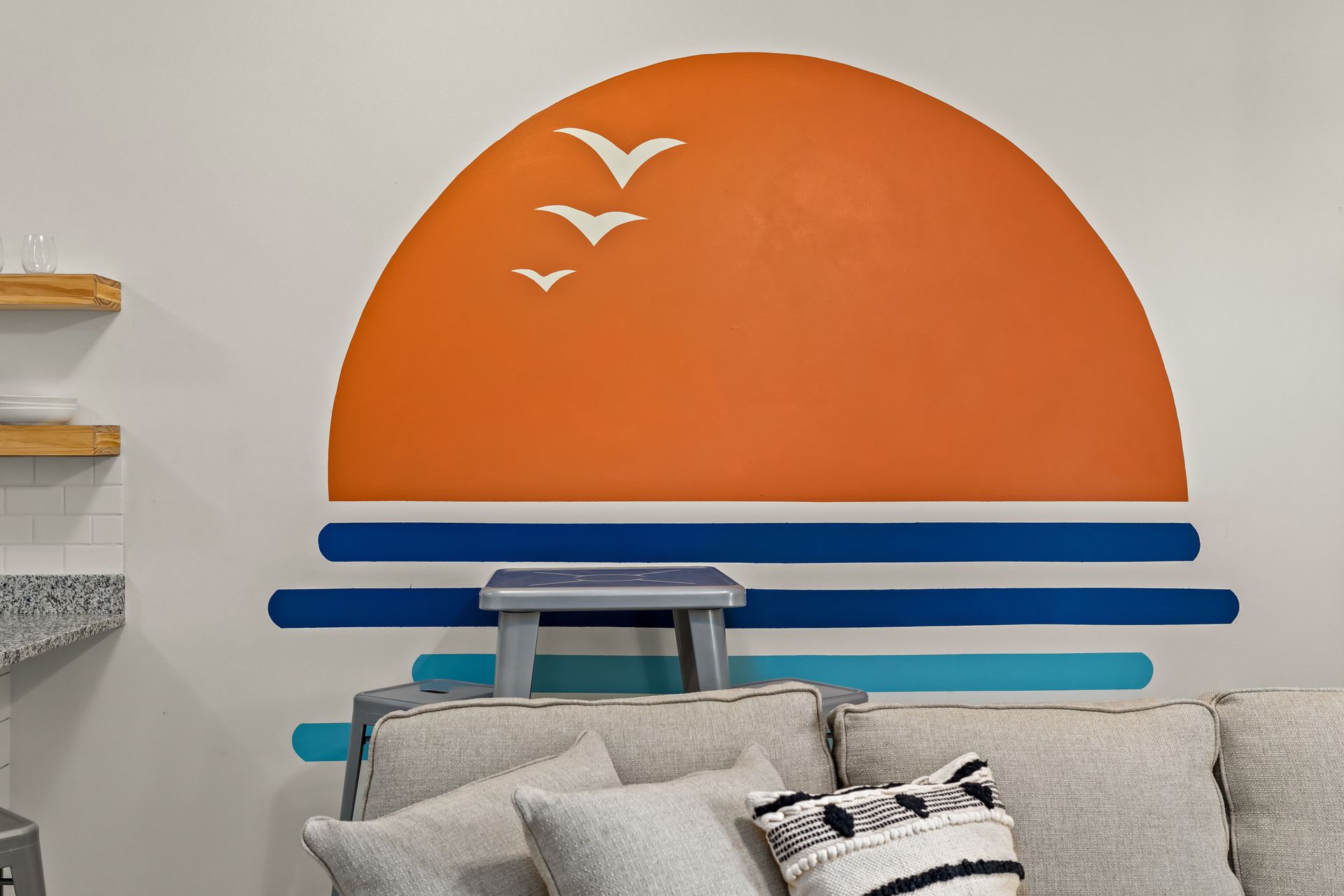 Orange sunset wall mural with white birds and blue stripes behind a gray sofa