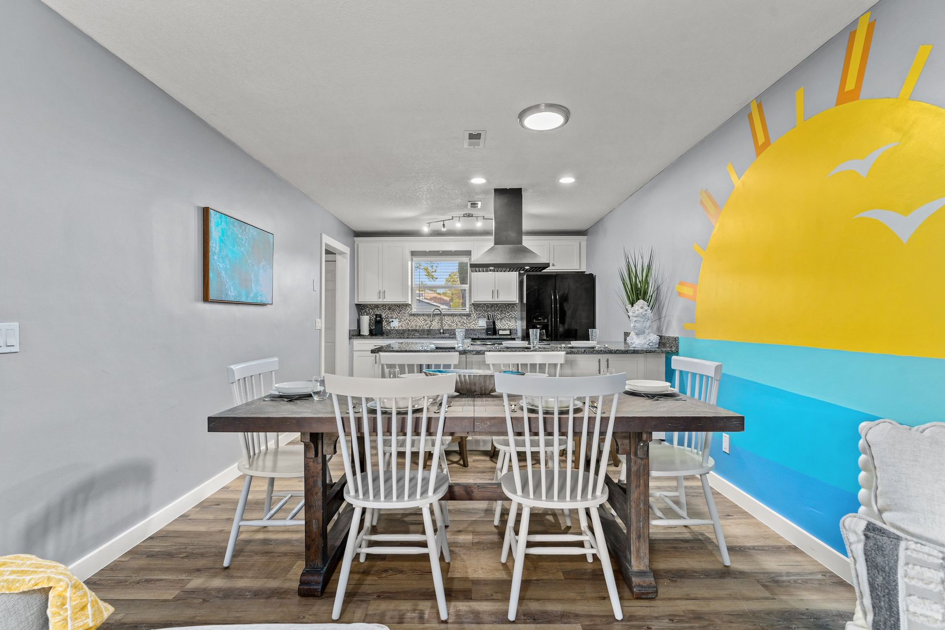 Bright dining room with white table and chairs, kitchen in back, and colorful sun mural on the wall