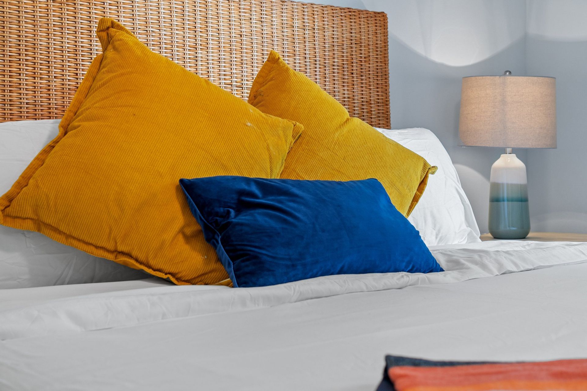 Bed with yellow and blue pillows against a woven headboard, beside a lamp and water bottle